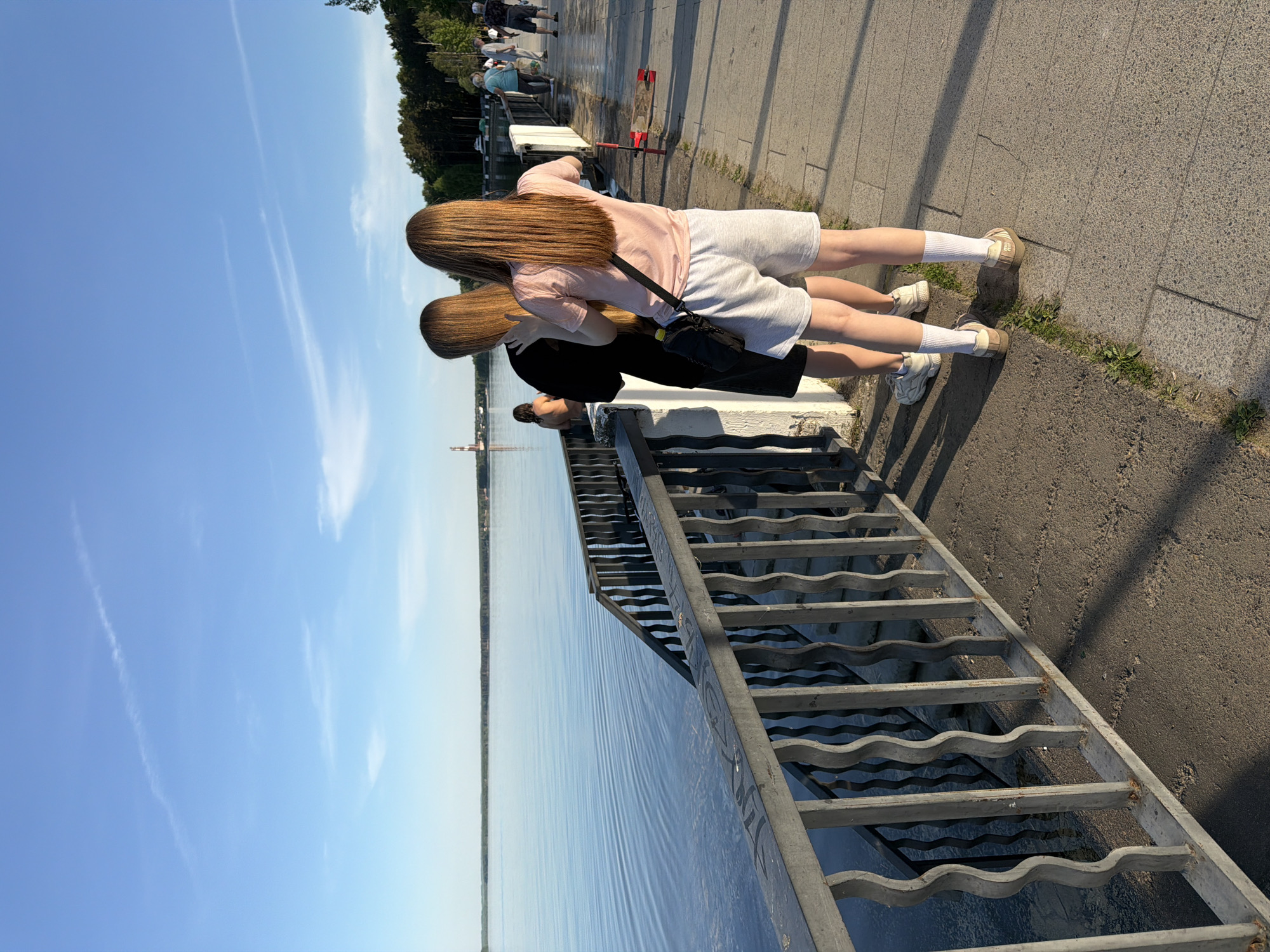 Two young women stand side-by-side on a concrete walkway, looking over a metal railing towards a body of water, possibly a river or lake. The scene is likely in the city of Chaikovsky, Russia. One woman wears a pink t-shirt and light grey shorts with a black shoulder bag, while the other wears a black t-shirt and darker shorts. Both are wearing white socks and sneakers. A metal staircase leads down to the water. The sky is blue with faint clouds. In the background, other people are walking along the walkway and trees line the edge of the waterway. A tower-like structure is visible across the water in the distance.