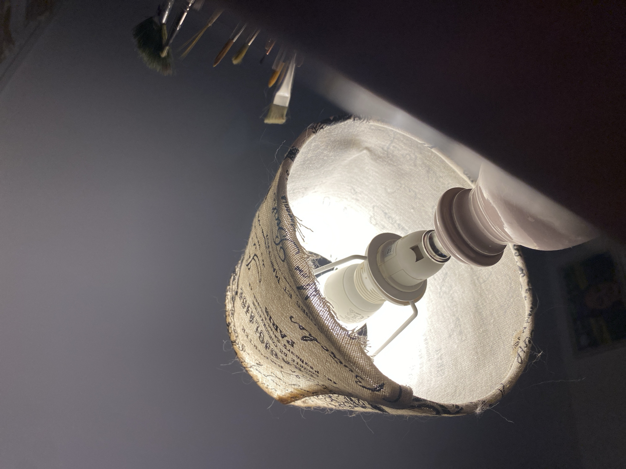 A low-angle, close-up shot focuses on a lamp with a textured lampshade adorned with text and a frayed edge. The lampshade is illuminated from within by a white LED bulb. The lamp base, a pale pink, is partially visible at the bottom. In the background, to the right, a collection of paintbrushes with green and yellow bristles is arranged, suggesting an artist's workspace. The overall environment appears to be indoors with dim, ambient lighting, casting shadows. The scene is devoid of any identifiable landmarks or elements that would suggest a specific location in Bílina, Czechia.