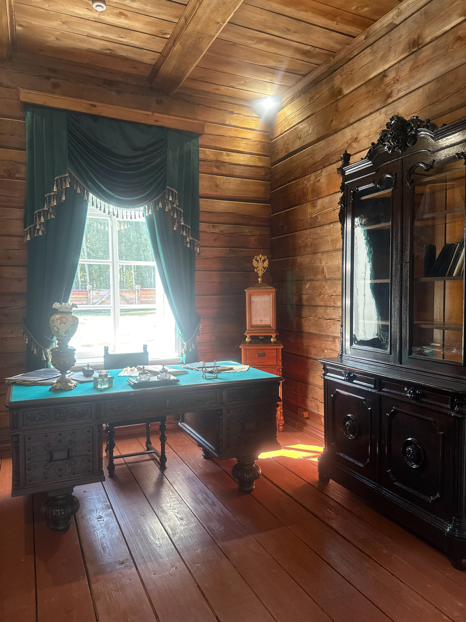 The image depicts a dimly lit, rustic interior of a wooden room, likely a study or office, within the "Taltsy" museum-reserve in Irkutsk, Russia. The walls and ceiling are constructed from rough-hewn horizontal wooden logs. In the center of the room, a large, ornate dark wood desk with carved detailing is positioned in front of a window. The desk is covered with a green felt cloth and displays a vintage lamp, an inkwell, papers, and other writing implements. Heavy emerald green velvet curtains with ornate tassels frame the window, which looks out onto a bright, sunlit outdoor scene with trees and a fence. To the right of the desk stands a tall, dark wood cabinet with glass doors, revealing shelves containing books. On the wall behind the desk, a small wooden stand with drawers holds a golden double-headed eagle emblem and a document. The floor is made of wide wooden planks with a reddish-brown finish, reflecting the sunlight streaming through the window. No people are present in the scene.