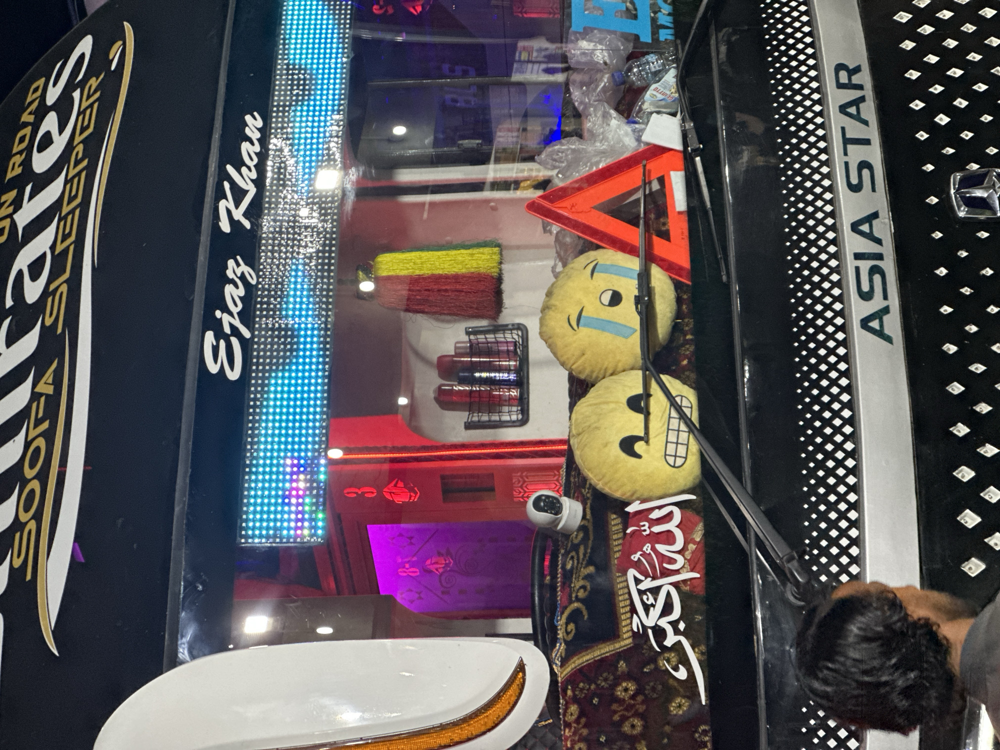 The image shows the front of a vehicle, likely a bus or van, potentially located in a city of Unknown, Unknown. The vehicle features lettering that says "Pirates SOFA SLEEPER," "Ejaz Khan," and "ASIA STAR." The windshield is adorned with two emoji pillows and a decorative Arabic script sticker. A windshield wiper is visible. Inside the vehicle, there is a red and yellow broom, a warning triangle, and various items including what appears to be plastic bottles and packaging. The interior is illuminated with LED lighting, including a strip of blue lights. A portion of a man's head is visible in the lower right corner.