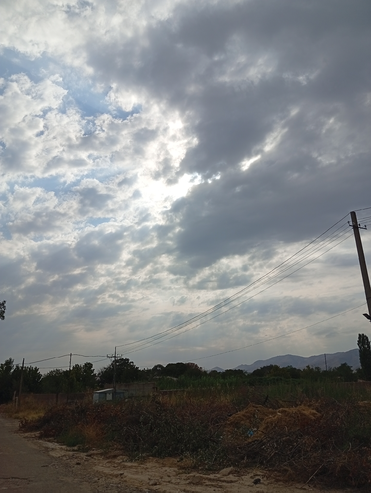 A landscape scene in Angren, Uzbekistan, depicts a partly cloudy sky with sunlight breaking through. In the foreground, a dirt road borders a patch of dry vegetation and debris. A utility pole with multiple wires stretches diagonally across the middle ground. Beyond this, a low wall separates the immediate area from a line of trees and a small building. In the distance, a range of mountains is visible under the cloudy sky.