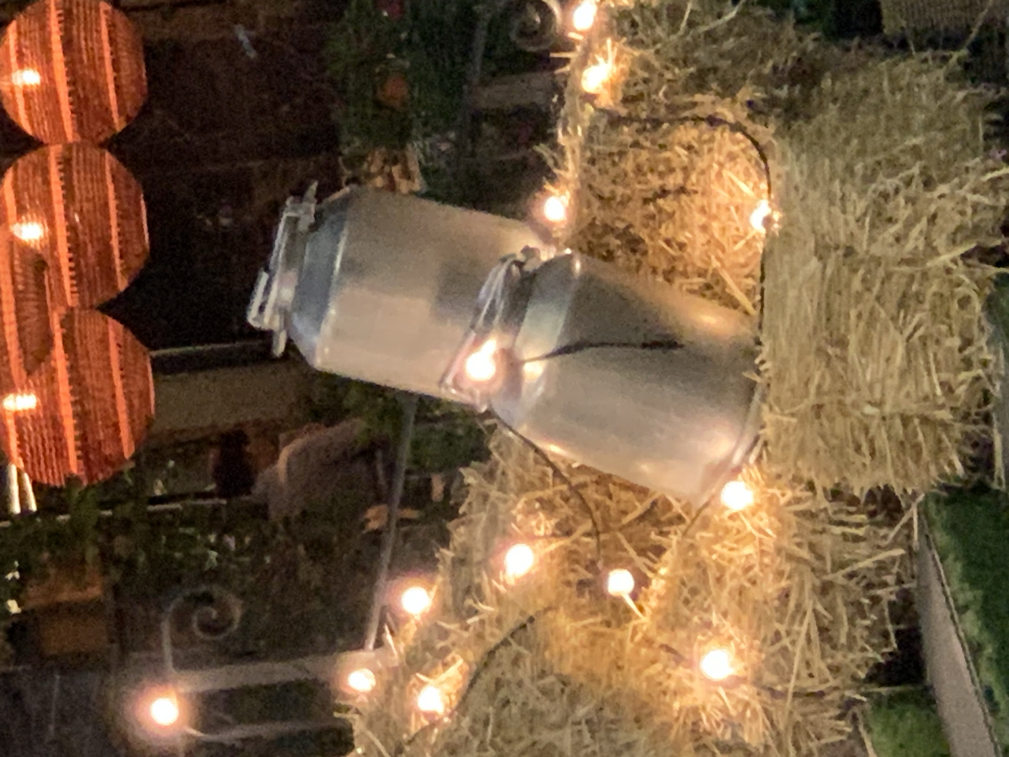 The image shows a close-up, low-light view of two metal milk cans stacked on top of each other, resting on bales of hay. A string of small, glowing lights is draped across the hay, illuminating the scene. In the background, there are out-of-focus elements including what appear to be decorative, woven light fixtures hanging from above, greenery, and a person in the distance wearing a light-colored shirt. The setting appears to be outdoors, possibly a decorative display or event.