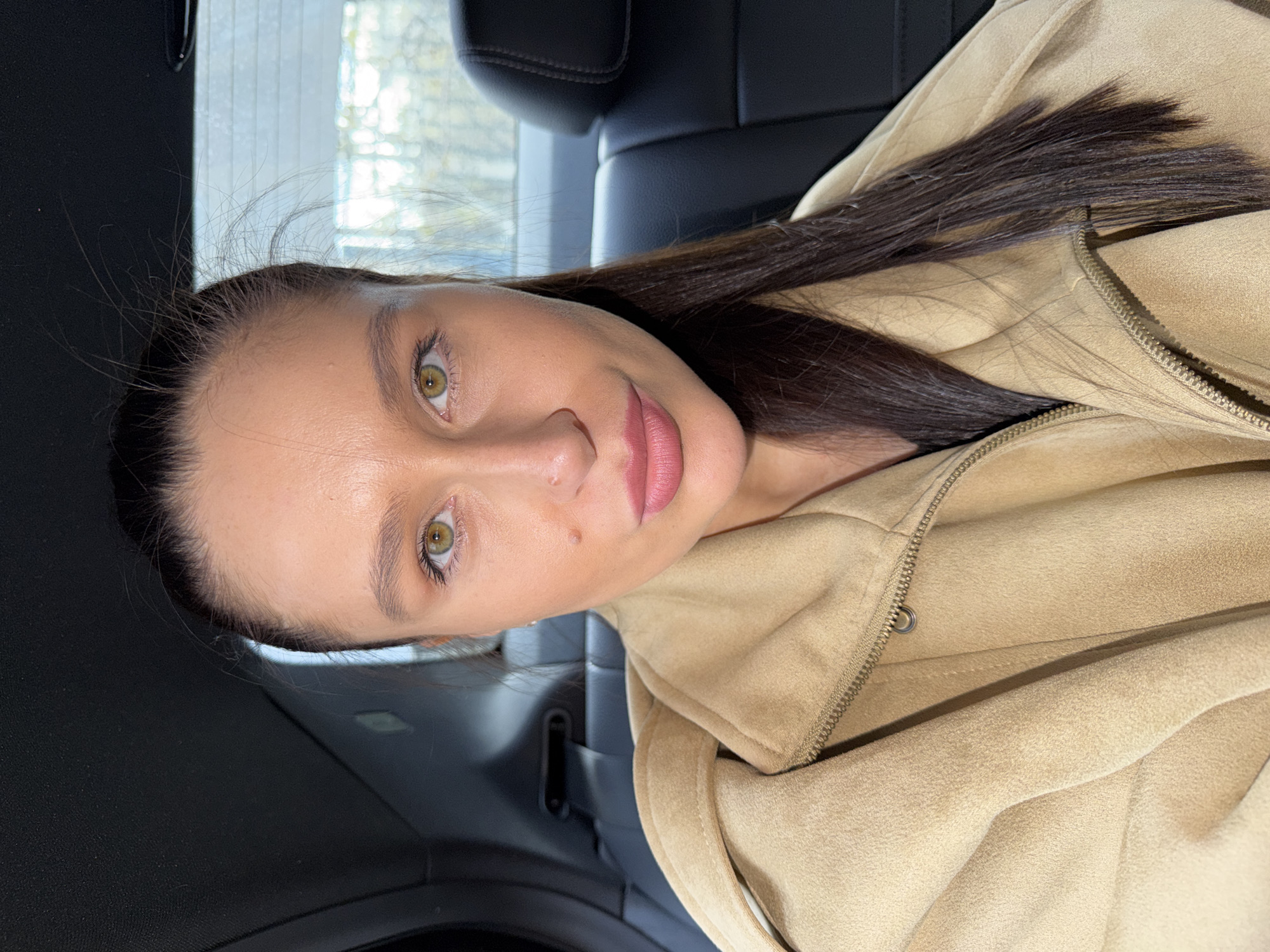 A woman is sitting in a car. She has long, straight, dark brown hair and fair skin. She is wearing a tan jacket. The background shows the car's interior and a blurred view of an urban environment. The location cannot be determined to be Khimki, Russia, based on this image alone.
