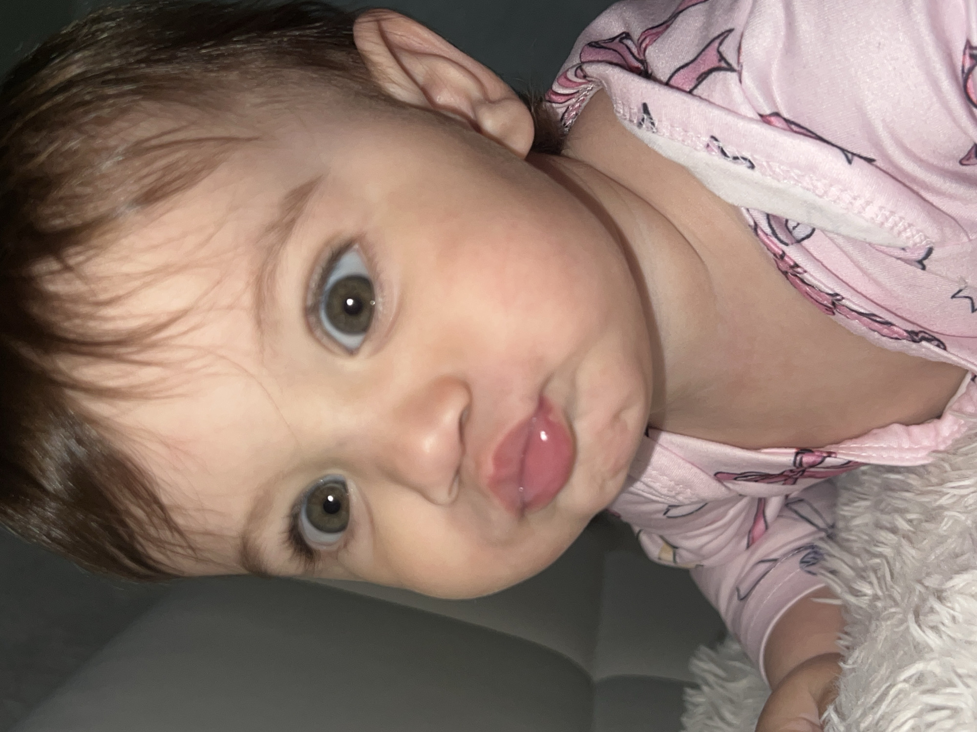 A close-up, slightly tilted, medium-shot photograph depicts a baby with light skin, brown hair, and hazel eyes. The baby is wearing pink pajamas with a unicorn pattern. The baby's lips are pursed in a pouting or kissing expression. The background includes a grey surface on the lower left and a fluffy white material on the lower right, which is touching the baby's arm. The location of Elena, Bulgaria, is not discernible from the provided image.