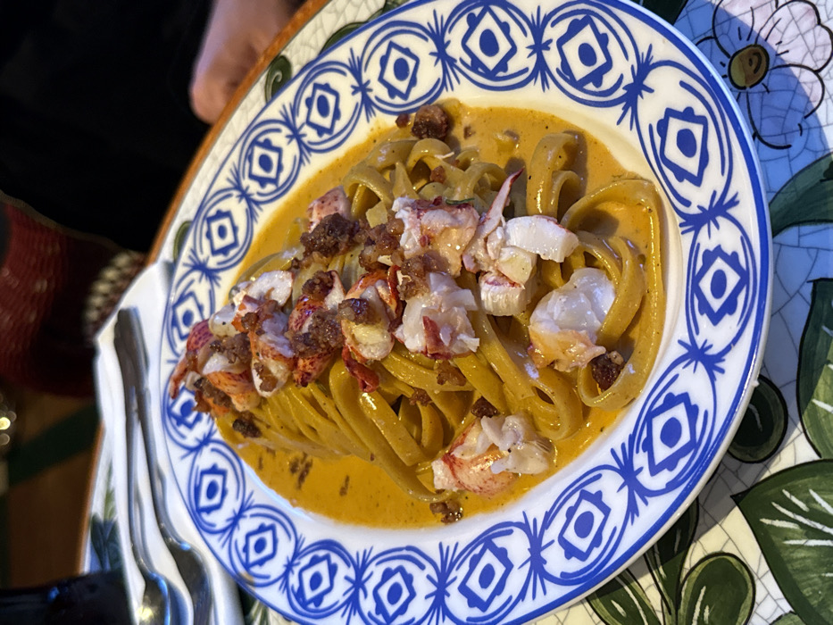 The scene depicts a plate of lobster pasta served in an indoor restaurant setting in Barcelona, Spain. The central subject is an oval white ceramic plate holding tagliatelle-style pasta coated in a creamy, orange-hued sauce. Generous portions of white lobster meat, flecked with red, are distributed atop the pasta, along with a dark, granular garnish, possibly crispy breadcrumbs or a savory crumble. The plate's wide rim features a repetitive blue and white pattern composed of geometric and stylized floral motifs. The plate rests on a table surfaced with green and white patterned tiles, forming a mosaic with abstract leaf or floral designs. To the upper left of the plate, a silver fork and spoon are neatly arranged on a folded white napkin, positioned next to a dark, cylindrical beverage bottle. The background is blurred, revealing textures suggestive of restaurant decor, including reddish-brown woven material, possibly from seating, and a dark, indistinct form to the right, consistent with another diner's presence. The lighting is warm and ambient, characteristic of an upscale dining establishment. The overall environment suggests a high-quality seafood restaurant, likely situated in a culinary district such as Eixample or near the waterfront in Barceloneta.