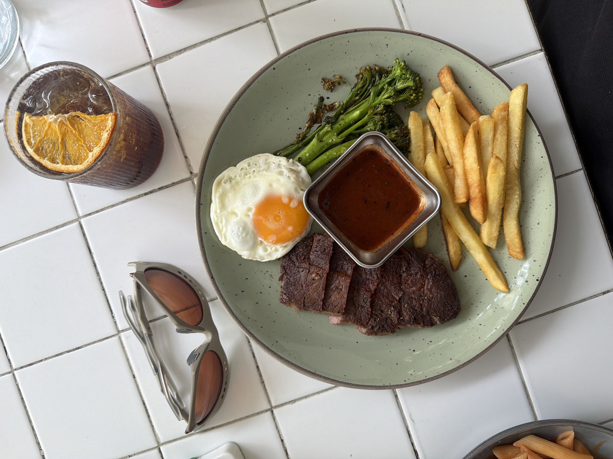 The image shows a meal and a drink on a white tiled surface. A plate contains steak, a fried egg, a ramekin of sauce, fries, and greens, likely broccolini. A glass beside the plate contains a dark beverage with ice and an orange slice. Sunglasses are placed next to the glass. A container with pasta is partially visible in the lower right. The location is presumed to be in Đà Nẵng, Vietnam, but there are no specific visual cues in the photo to confirm.