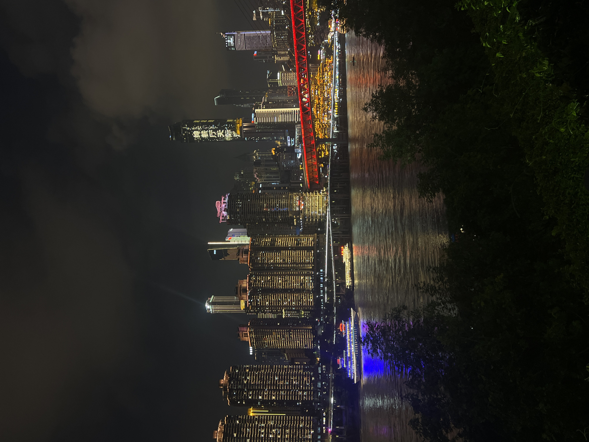This is a nighttime, horizontal, eye-level shot of the Yangtze River in Chongqing, China, specifically near Jiangbeicheng Subdistrict. The scene is dominated by the illuminated cityscape on the far bank, with numerous tall buildings featuring a warm, golden glow from their windows. A prominent red bridge spans the river, its structure illuminated with red lights. Several tour boats with colorful lights are visible on the water, with one in the foreground reflecting blue light. The foreground is a dark area with foliage, suggesting the viewer is on a higher vantage point looking down. The sky is dark and cloudy, with some faint lights visible behind the clouds.
