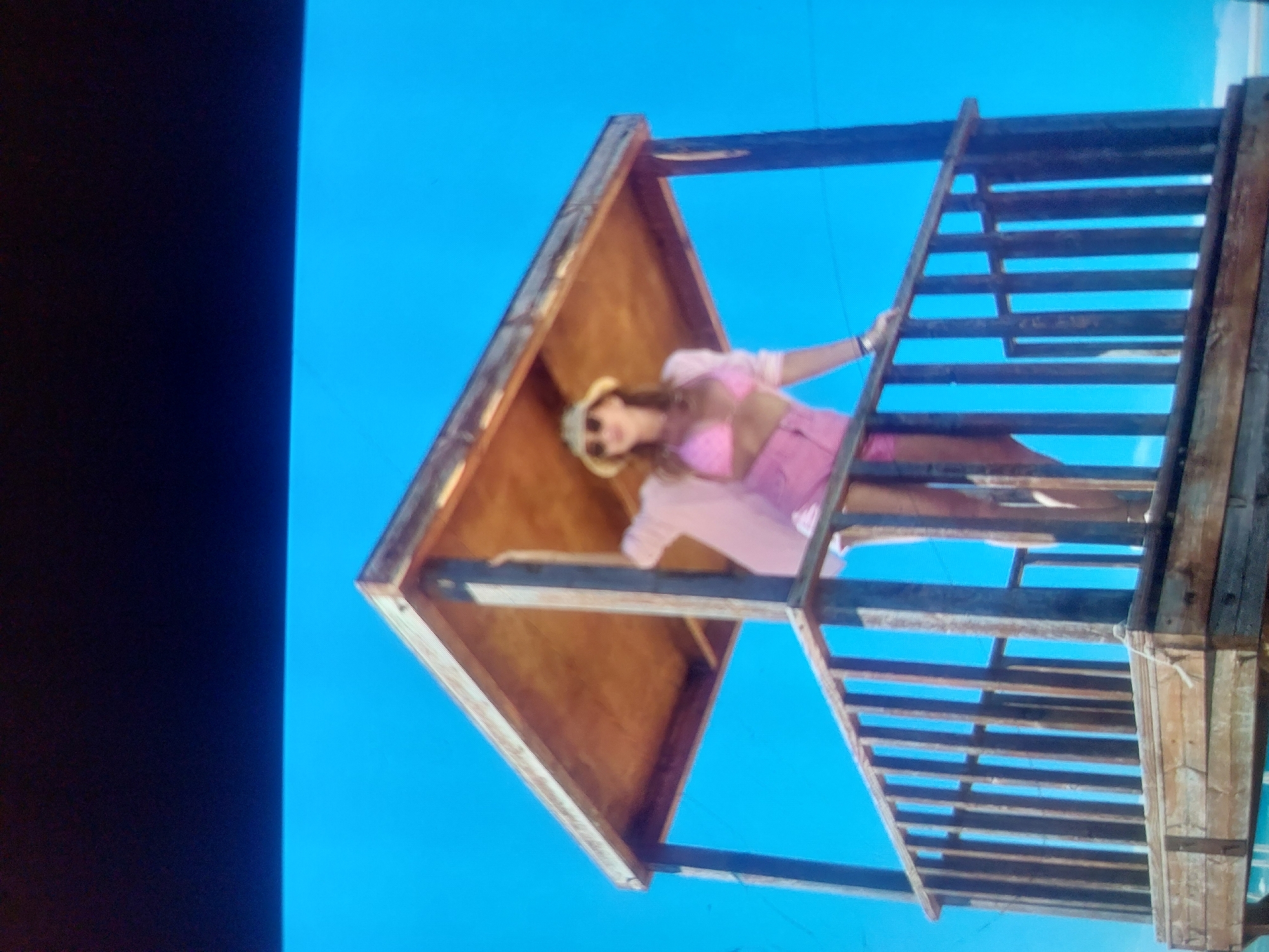 A low-angle shot depicts a woman standing in the upper portion of a wooden structure with a ladder, set against a bright blue sky. The woman is wearing a pink outfit, sunglasses, and a hat. The wooden structure consists of a ladder and a box-like enclosure above it. The image appears to have been taken in Tsarevo, Bulgaria.