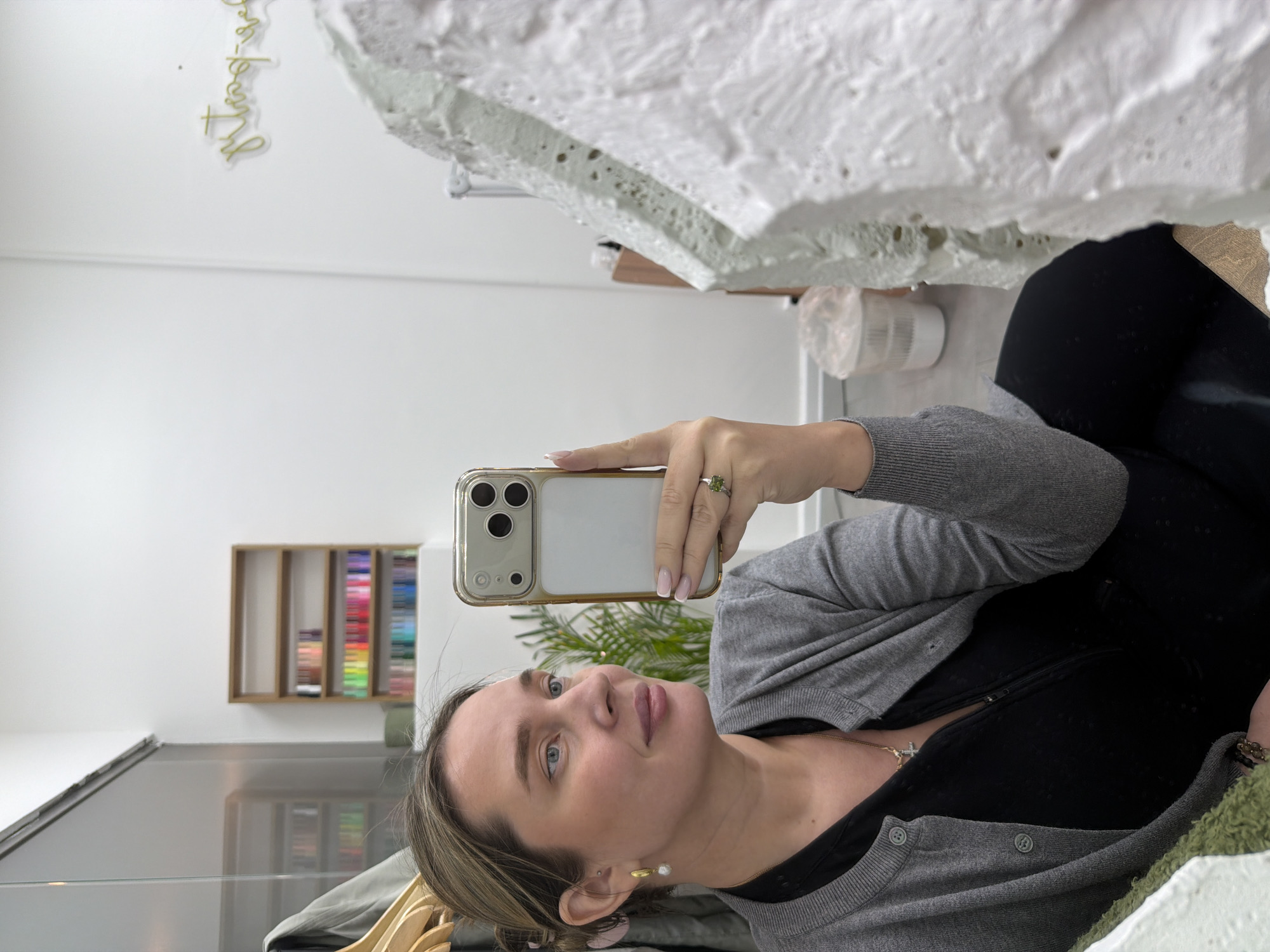 A woman is captured in a selfie, holding a gold-cased smartphone in her right hand. She has fair skin, blue eyes, and light brown hair, and is wearing a grey cardigan over a black top. Her jewelry includes a green stone ring, a gold cross necklace, and gold and pearl drop earrings. Her fingernails have a light pink base with white French tips. She looks calmly towards the camera with a neutral expression.
The setting appears to be a modern, well-lit indoor space, possibly a studio or salon, characterized by white walls and a reflective grey floor that shows a blurred reflection of the room, including some green foliage. In the background, a wooden shelf unit against the wall displays rows of colorful items, likely art supplies or products. Prominently featured in the upper right is a large, textured white object with a porous, uneven surface, possibly a decorative art piece or architectural element. A small white cylindrical object rests on a surface below it. On the white wall in the upper left, a gold script sign is partially visible, reading "It's always the..." with the final word indistinct due to styling or blur. The image depicts an indoor scene in Moscow, Russia.