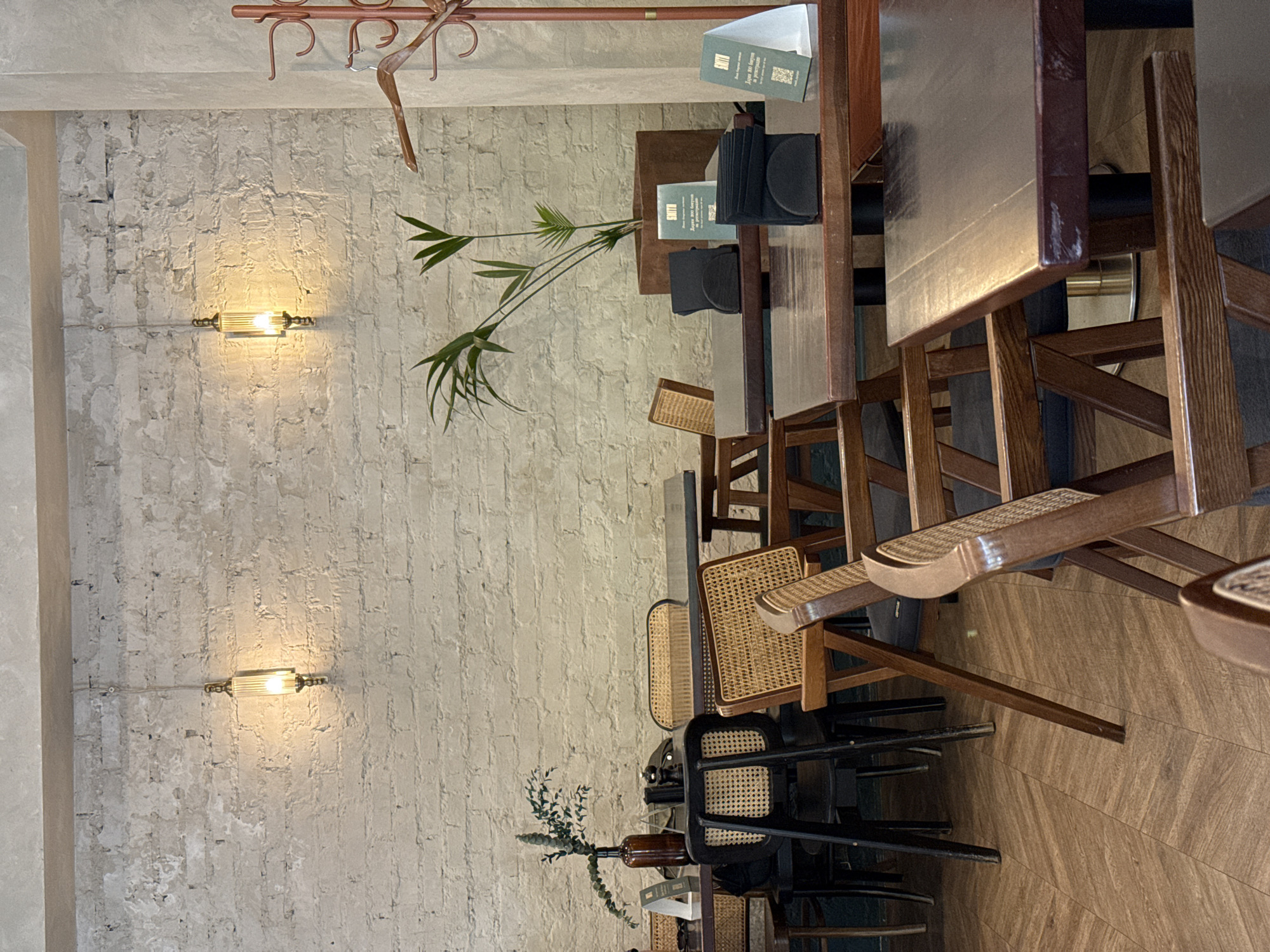 The image captures the interior of a dining space, likely a cafe or restaurant. The scene is dominated by wooden tables and chairs, exhibiting a blend of modern and rustic styles. The chairs have woven wicker seats. A narrow rectangular table is visible, with some greenery/plant as a table decoration.
The walls are rough and textured, painted in a light, neutral color, possibly white or a very light gray. Two sconce lights are spaced along the wall. A coat rack is suspended near the top of the image frame.
The overall environment suggests a contemporary interior with natural elements and neutral tones. This style and aesthetic is consistent with cafes found in various cities around the world, including Krasnoyarsk, Russia. However, without further information or context, definitively determining the specific location within Krasnoyarsk is not possible based on the image alone.