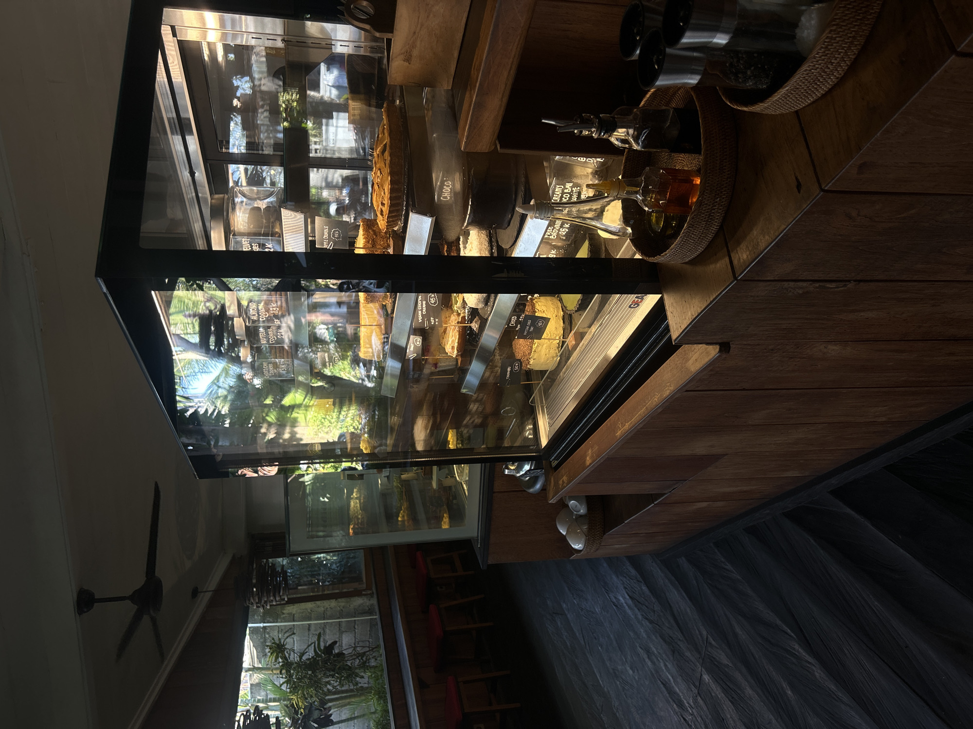 The image shows the interior of a cafe in North Kuta, Indonesia. A large refrigerated display case filled with various cakes and pastries is prominently featured in the center of the frame. The display case has glass doors and is lit from within, illuminating the baked goods. On the counter next to the display case, there are several bottles of oil and vinegar in a woven basket, along with salt and pepper grinders. To the left of the display case, there is a seating area with red stools and a window overlooking an outdoor area with plants. The floor is dark, with a herringbone pattern. A ceiling fan is visible in the upper left corner of the image. The overall atmosphere is dimly lit, with natural light filtering in from the window.
