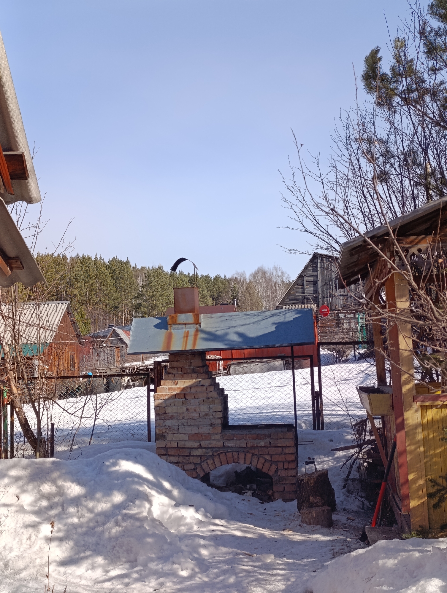The image shows a snowy backyard scene in what appears to be a rural or suburban area. In the foreground, there is a brick outdoor grill with a metal roof and chimney. A pile of snow sits in front of the grill. To the right of the grill is a wooden structure with a yellow wall. An axe is stuck in a stump of wood near the grill. Behind the grill, a chain-link fence separates the yard from neighboring properties. In the background, there are several wooden buildings and a forest. The sky is clear and blue.
Based on the visual characteristics of the buildings and landscape, this could be a location near Krasnoyarsk, Russia.