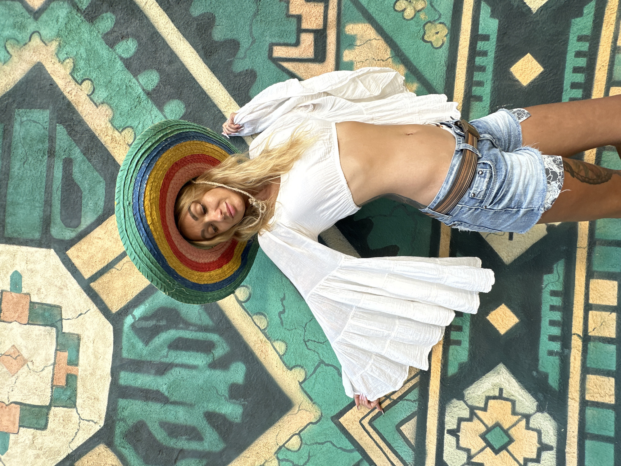 A woman with blonde hair and closed eyes, wearing a colorful sombrero, a white crop top with bell sleeves, and denim shorts, poses in front of a mural on a wall. The mural features geometric patterns in green, yellow, and tan. The scene is set in Mui Ne, Vietnam, likely on a street or public area. The woman's left arm is extended to her side, and she holds the fabric of her top in her right hand. Tattoos are visible on her legs.