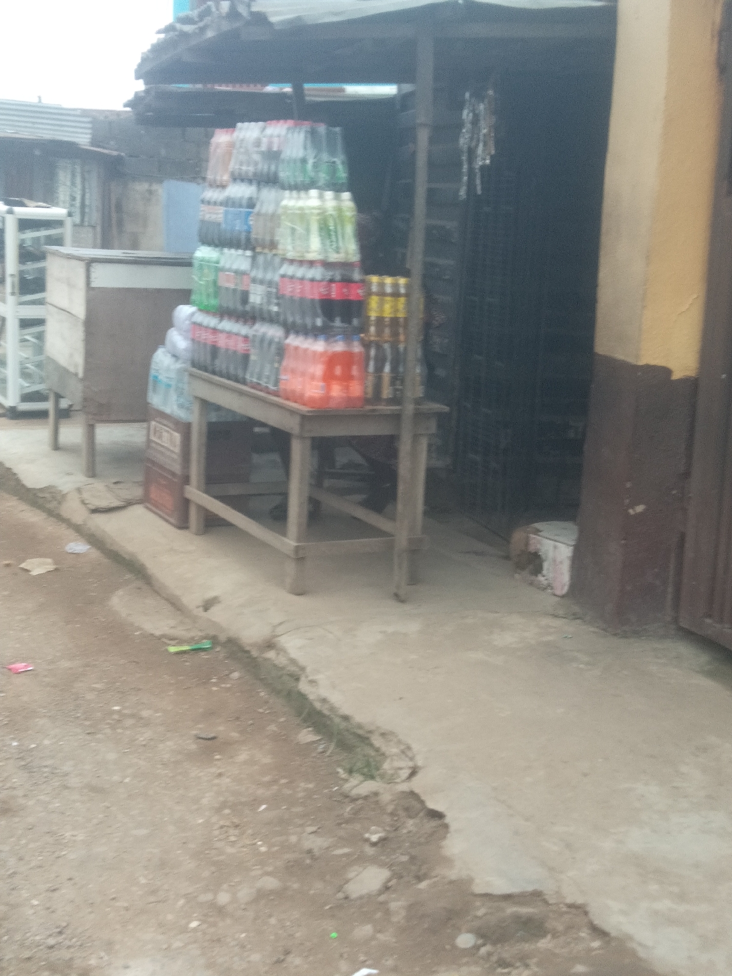 The image depicts an outdoor market stall in Shangisha, Nigeria. The stall is stocked with numerous bottles of beverages, primarily soft drinks, arranged on a wooden table and on the ground. The bottles are predominantly in plastic packaging and are stacked in various rows and columns. Some of the bottles appear to be clear, while others contain colored liquids, suggesting different flavors or brands. In the background, there is a building with a corrugated metal roof and walls, typical of market structures in the area. The ground in front of the stall is a dirt and concrete surface, with some debris scattered around. The overall scene suggests a commercial transaction is taking place or has recently occurred, with goods displayed for sale. There are no people clearly visible in the image, and the focus is on the merchandise and the stall setup.