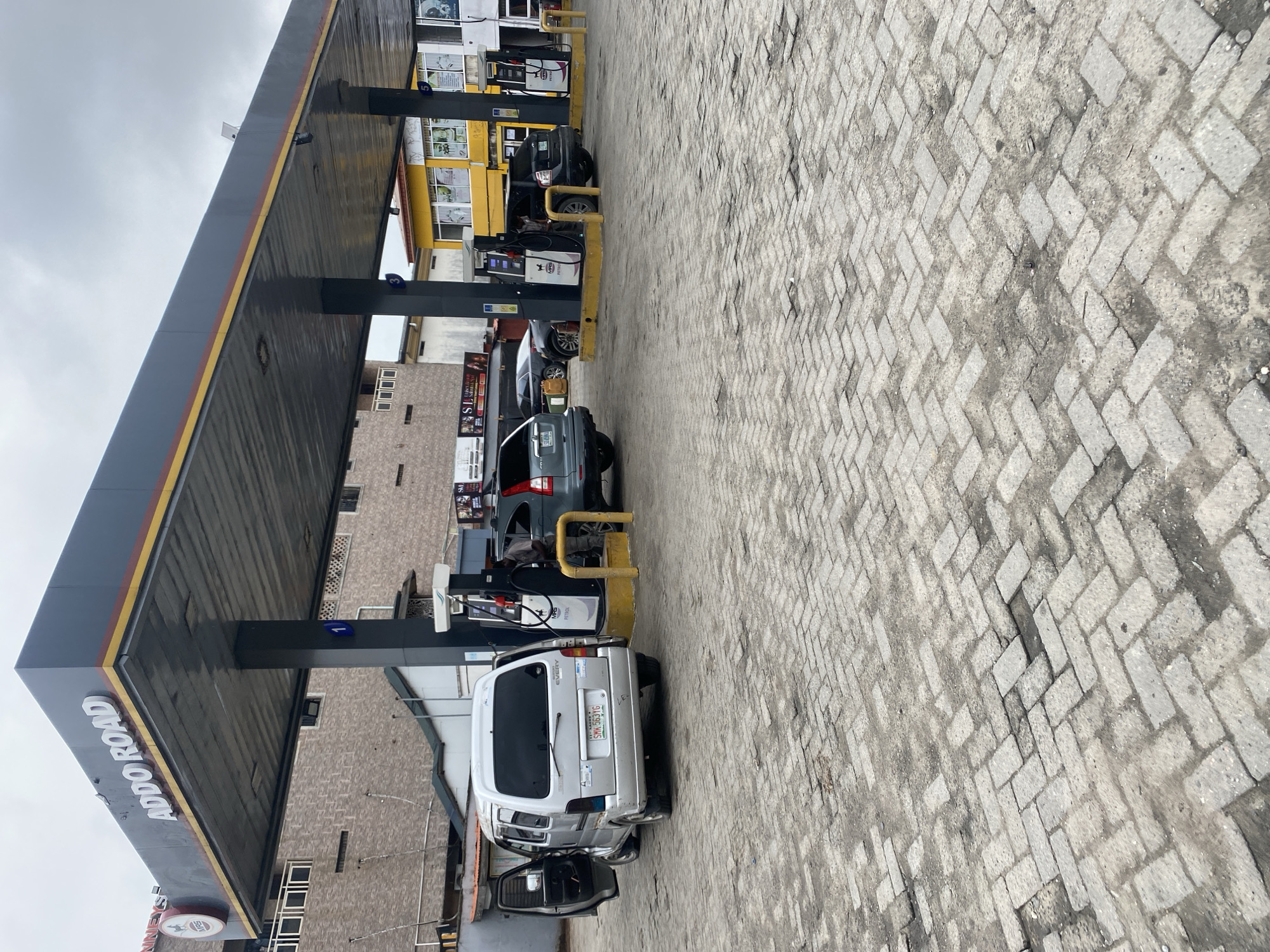 An outdoor, low-angle shot shows the forecourt of a gas station, identified by the sign "ADDO ROAD" on the canopy. The ground is paved with grey interlocking bricks. To the left, a silver van is parked with its driver's side door open, and a fuel nozzle is inserted into its gas tank. Next to the van, a dark grey SUV is parked, and a person in a grey shirt is standing by a fuel pump. Further to the right, another dark grey SUV is parked at a fuel pump, and a person in a dark shirt appears to be operating the pump. In the background, a brick building with multiple windows stands behind the gas station canopy, and a yellow building with windows is visible to the right. The sky is overcast and grey.