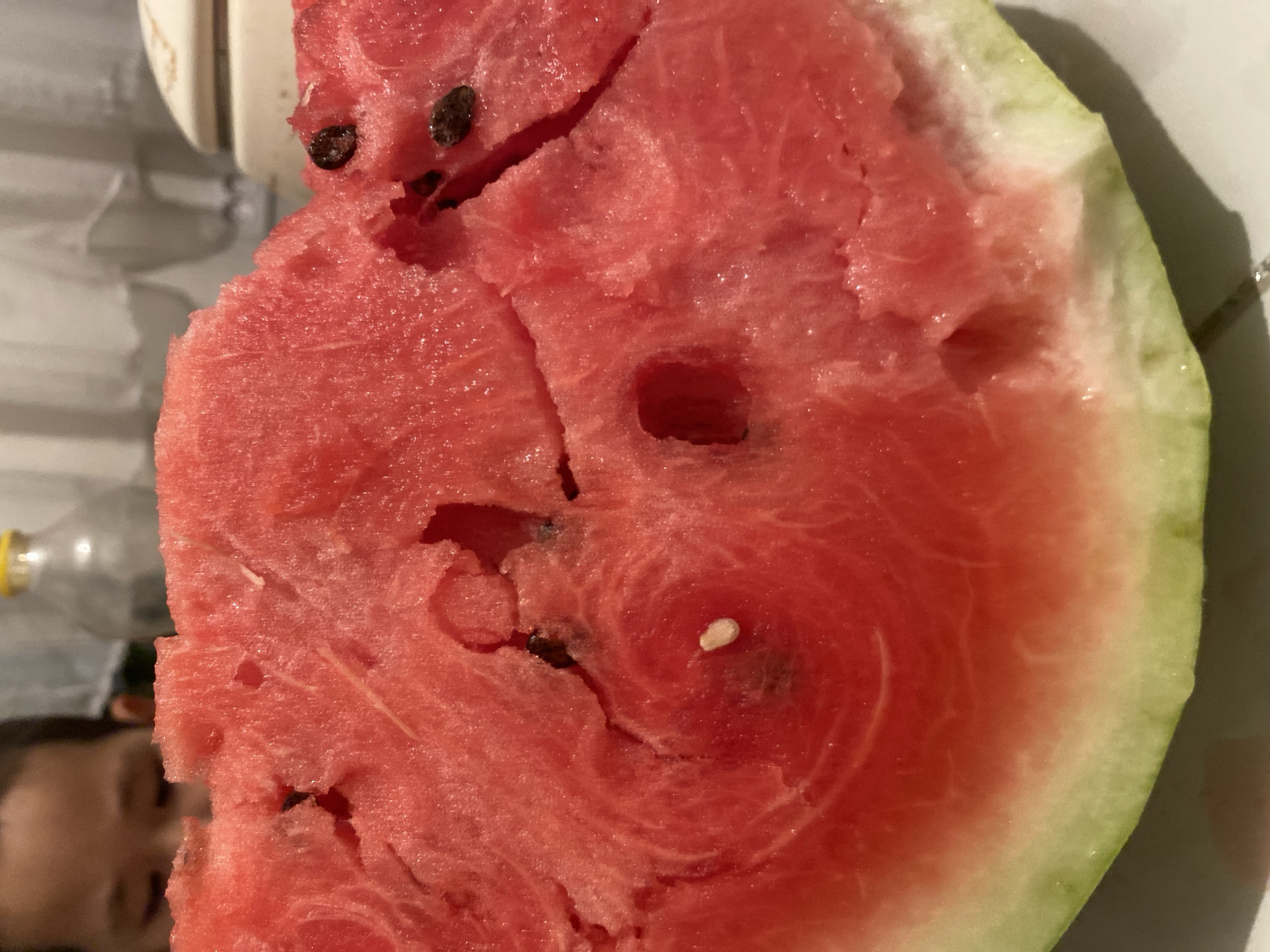 In the foreground of the image, a large, juicy slice of watermelon is prominently displayed. The watermelon flesh is a vibrant red, with visible fibrous strands and several dark seeds. A single white seed is also noticeable. The rind is a pale green transitioning to white near the flesh. The watermelon is positioned on a white surface, likely a plate or table. In the background, out of focus, a child's face is partially visible, looking down towards the watermelon. Further in the background, indistinct objects and white curtains suggest an indoor setting. The scene appears to be in a residential environment, possibly a home. There is no information available to specify the location within Boraldai, Kazakhstan.