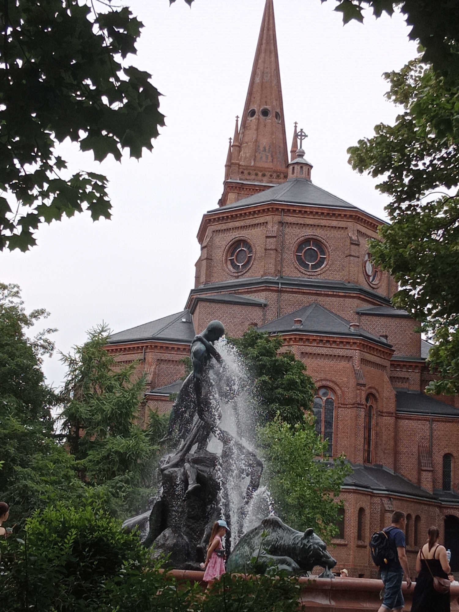 The image captures a fountain sculpture in the foreground, with a large brick church as the backdrop. The fountain features a dark bronze statue of a man in motion, surrounded by splashing water. In front of the man, a bronze sculpture of a bear lies with its head down, appearing to drink from the water. To the right of the fountain, two people are walking past, a man with a backpack and a woman in a black dress. To the left of the fountain, a young girl in a pink dress stands facing away from the viewer, looking at the fountain. Behind the fountain, a large, multi-tiered brick church dominates the scene, with a tall, pointed steeple and multiple arched windows. Lush green trees and foliage surround the fountain and partially obscure the church. The sky is overcast, suggesting a cloudy day. This fountain is located in Bydgoszcz, Poland.