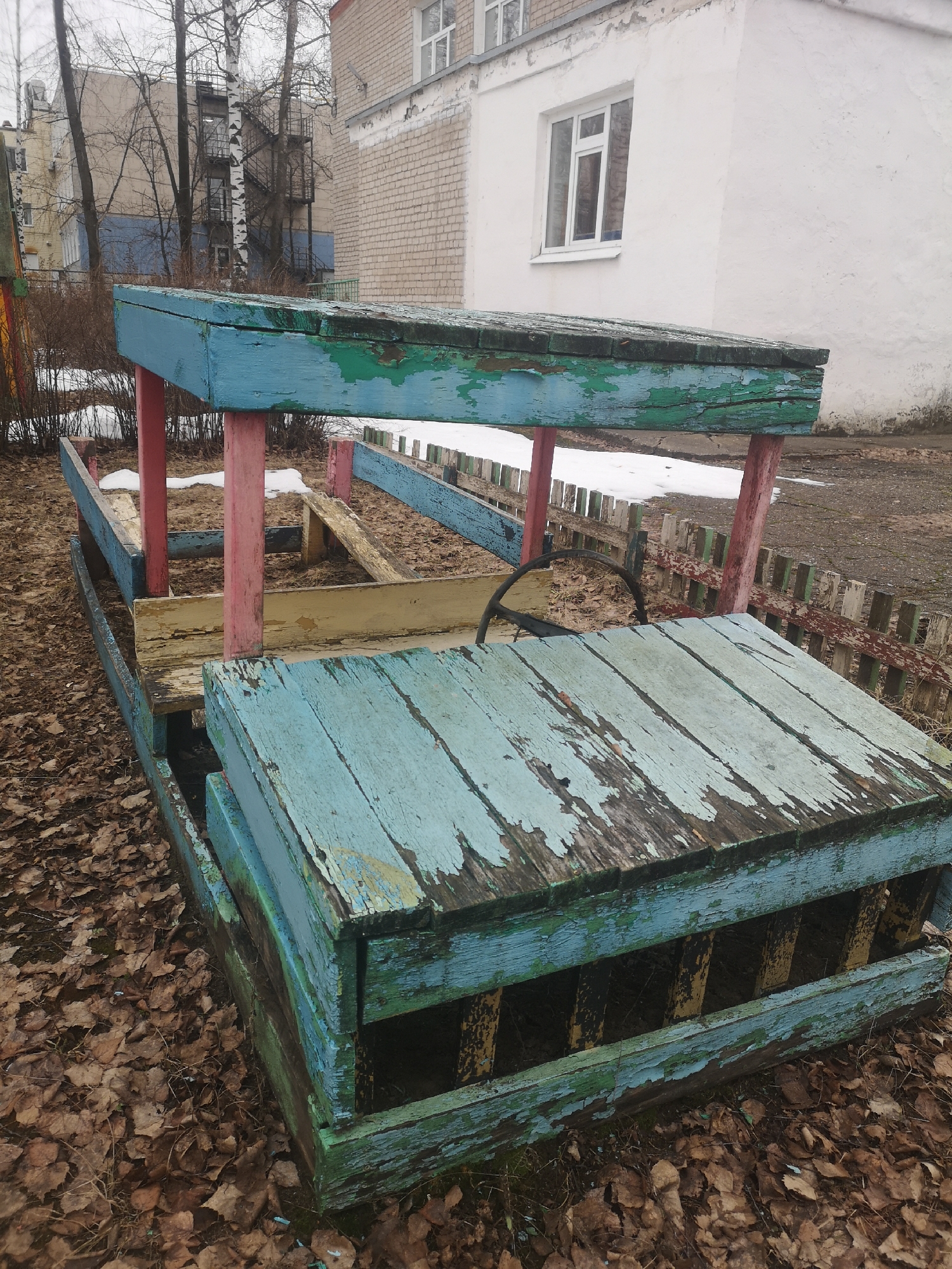 The image depicts an aged, wooden playground structure in the form of a boat, situated in what appears to be a park or play area. The location is in Chaikovsky, Russia. The structure has a weathered blue and green paint job with chipping and peeling paint. The supporting posts are pink. The surroundings consist of fallen leaves on the ground, suggesting it is either late autumn or early spring. In the background, there are residential buildings and trees. There are small patches of snow visible, indicating a cold climate. A short wooden fence is partially visible in the background.