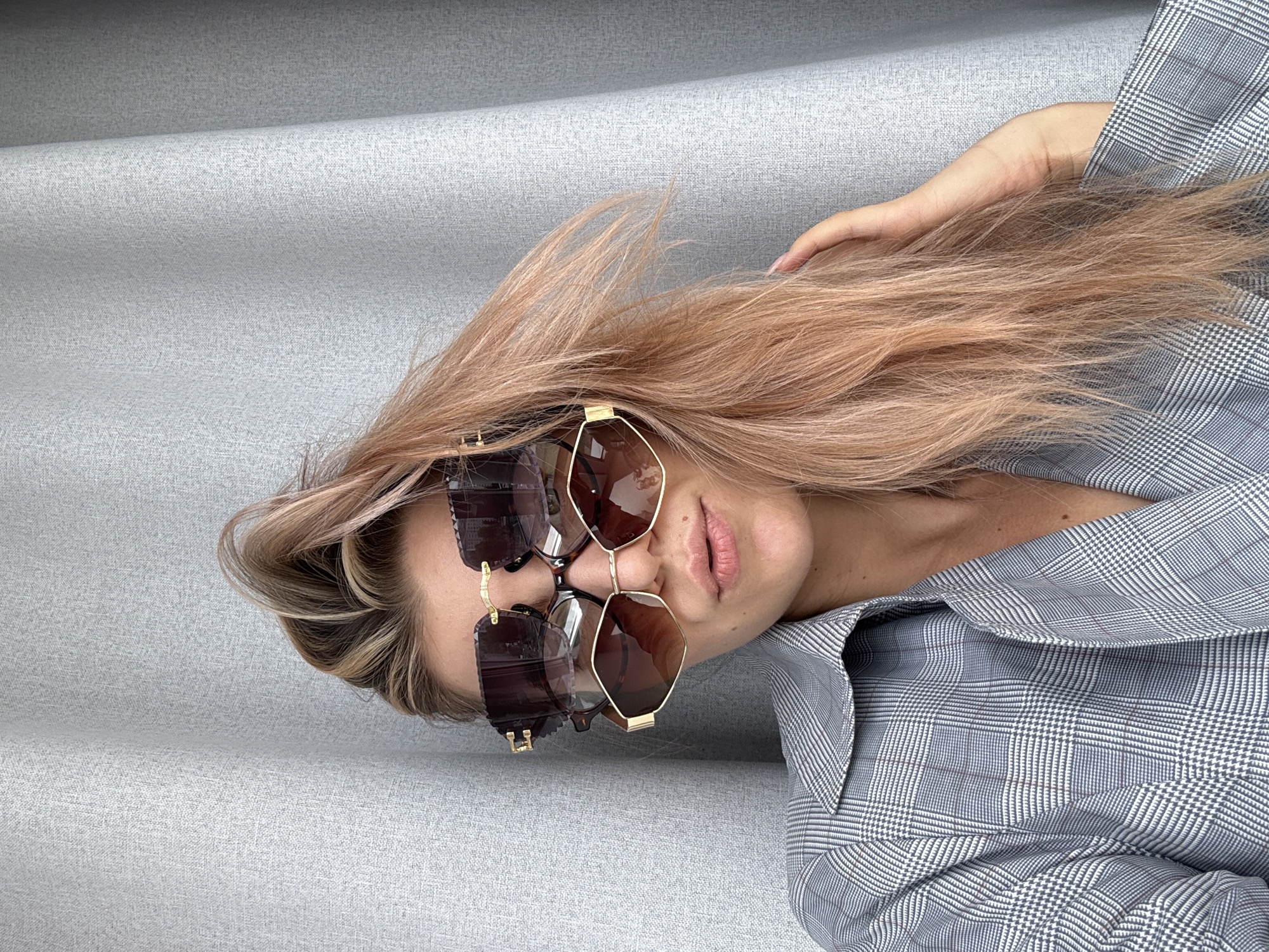 A woman with blonde hair and light skin is lying on a gray, textured surface, possibly upholstery. She is wearing sunglasses with gold frames and brown lenses. Her lips are pink, and she is wearing a gray and white plaid shirt. Her left hand is placed in her hair. The location is unknown.