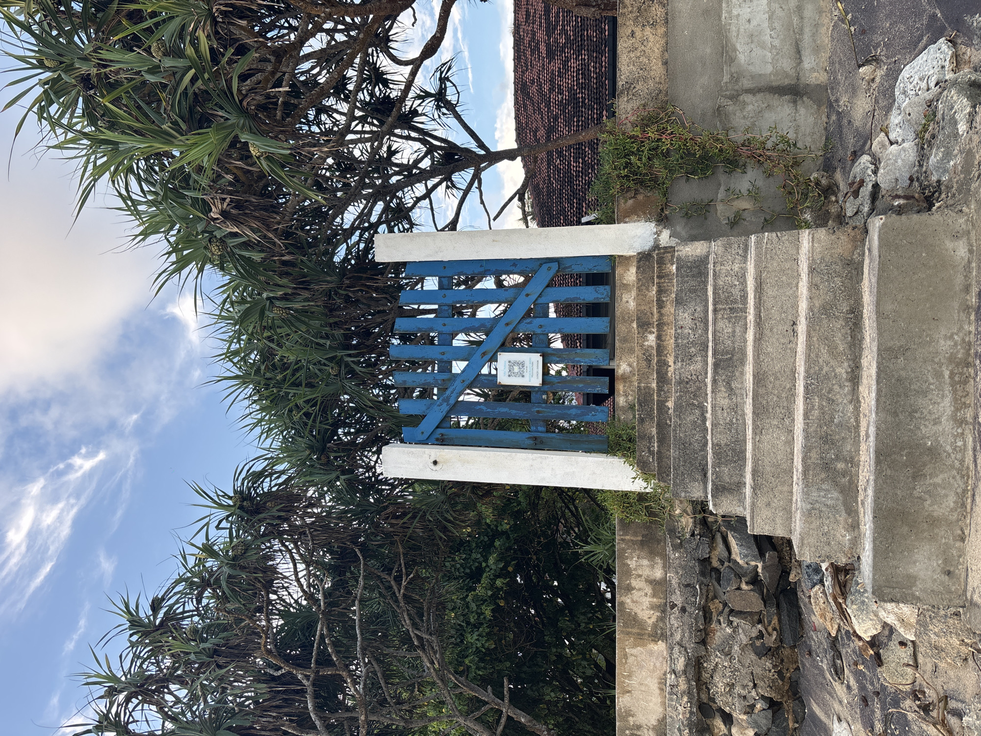 A blue wooden gate blocks the top of a set of concrete stairs leading up to a tiled-roof building in Thalaramba, Sri Lanka. The gate is flanked by white concrete posts. A QR code is affixed to the gate. Dense tropical foliage surrounds the structure, and a blue sky with wispy clouds is visible above. The concrete stairs are weathered and partially overgrown with greenery. Rocks are visible along the side of the staircase.