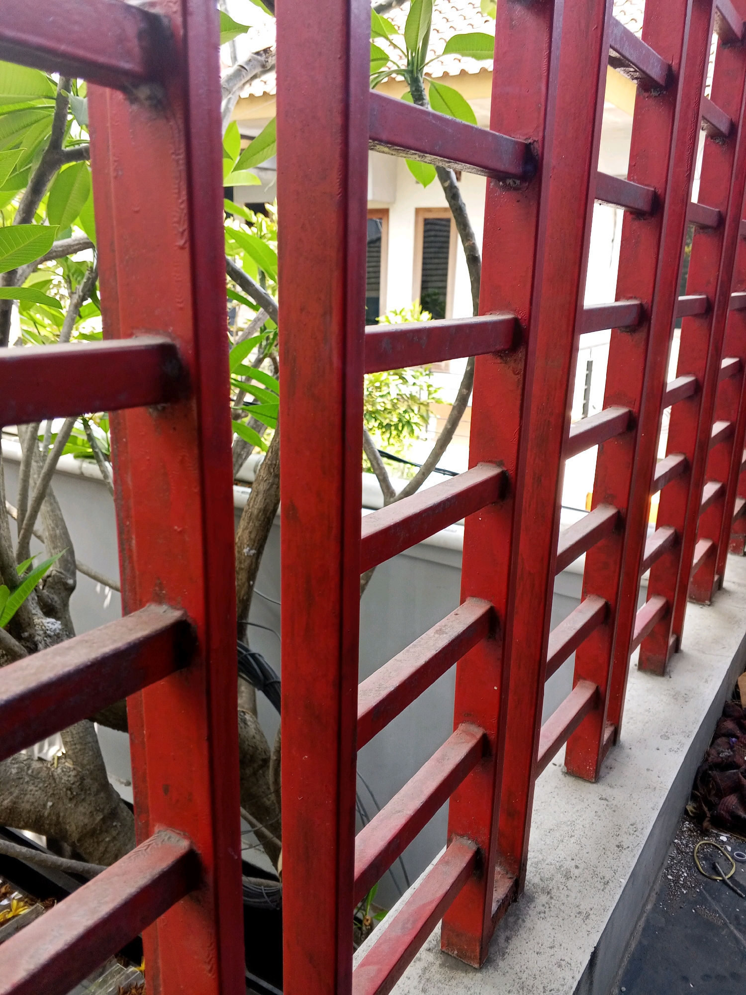 The image depicts a close-up view of a red metal fence with vertical bars and horizontal supports. Behind the fence, there are green leaves and branches of a tree, suggesting an outdoor setting. A building with a white wall and windows is partially visible in the background. The fence appears to be weathered, with some rust or discoloration visible on the red paint. The fence is situated on a concrete ledge. The scene is consistent with a residential or commercial area in Condongcatur, Indonesia, possibly a garden or the perimeter of a property. There are no people visible in the image. The lighting suggests it is daytime.