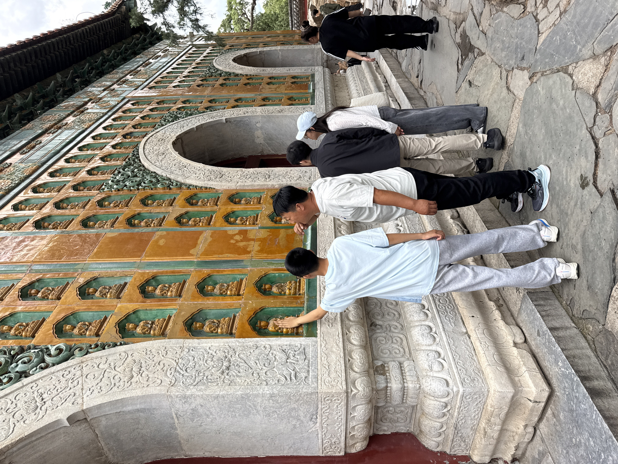The image depicts an outdoor scene at the Hall of Ten Thousand Buddhas (Wanshou Mountain) within the Summer Palace (Yiheyuan) in Haidian District, Beijing, China.
A large, ornate building facade dominates the scene, characterized by its extensive wall decorated with a grid of recessed niches. Each niche, framed in green glaze and set against an orange-glazed tile background, houses a small, golden-colored, seated Buddha figurine. Three large, grey stone arched doorways are visible along the wall; their surrounds and the building's base are intricately carved with floral and scrollwork patterns. Decorative green elements are present above the arches and along the upper section of the wall, beneath a dark-tiled roof with extended eaves visible at the top right.
In the foreground, a young male, appearing to be between 8 and 12 years old, wearing a light blue t-shirt and grey sweatpants, stands with his back partially to the viewer, reaching his right hand to touch a Buddha figurine in a lower niche. To his immediate right, an adult male in a white t-shirt and dark pants observes the wall, looking upwards with his mouth slightly open. Further back, a male in a black jacket and tan pants stands next to a female wearing a white long-sleeved shirt, blue jeans, and a light blue baseball cap, both also looking up at the building. In the background on the right, an adult female in a black tracksuit walks away from the viewer along a stone path.
The ground is paved with irregularly shaped grey stone slabs, displaying cracks and uneven surfaces. Trees are visible in the background, and the sky appears overcast.