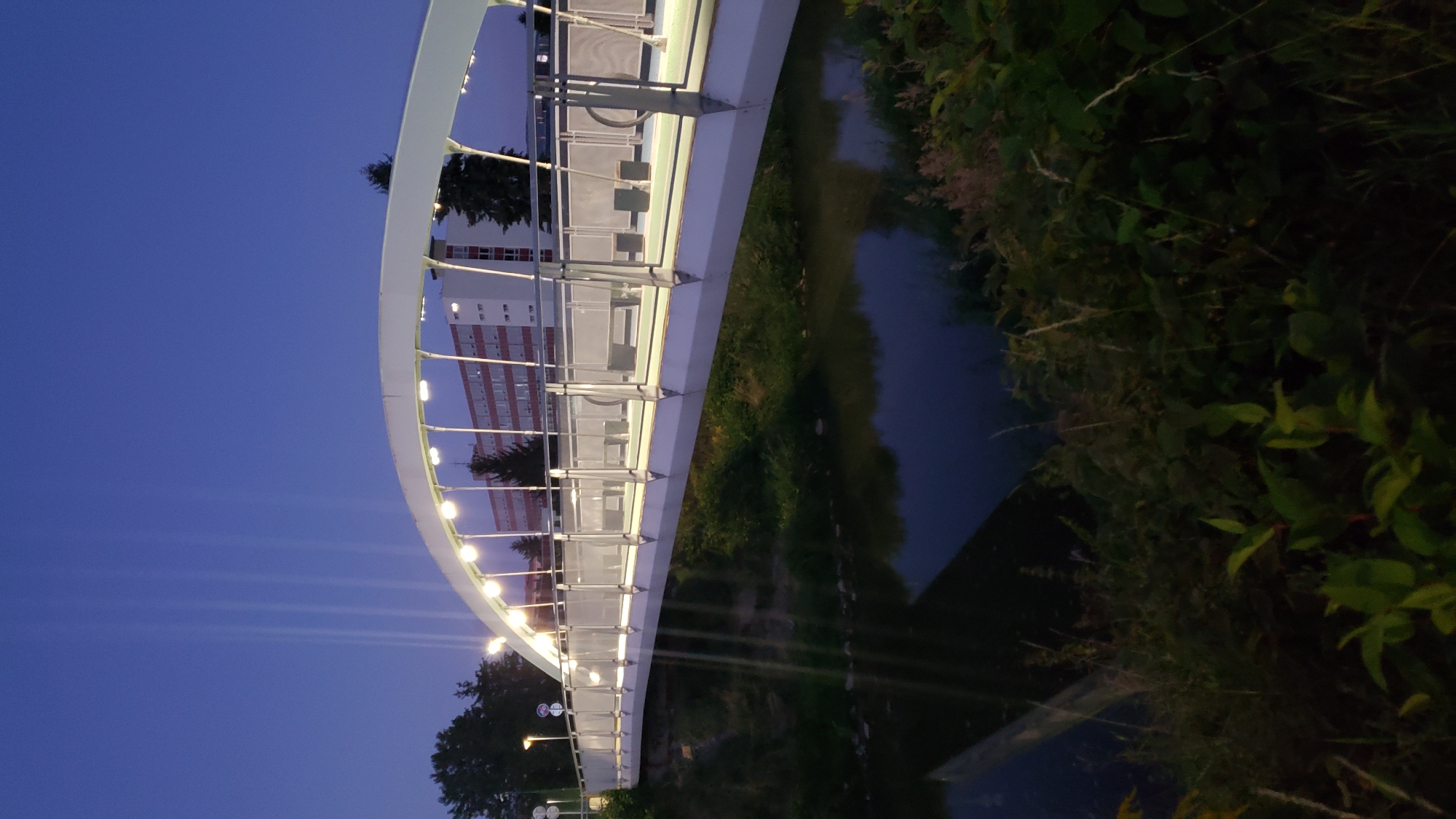 This is a vertical, medium shot photograph taken at dusk of the footbridge over the Sekčov stream in Prešov, Slovakia. The bridge is modern and white with a curved superstructure illuminated by a series of lights. It has a metal railing with glass panels. Along the bridge's walkway, there are several benches and seating areas. The Sekčov stream flows beneath the bridge, reflecting the lights from the bridge. The banks of the stream are covered in lush green vegetation. In the background, a multi-story building with a red facade and white window frames is visible. Tall trees are also present on the left side of the image. The sky is a deep blue, characteristic of twilight. Several vertical light beams are visible in the sky above the bridge, suggesting a light installation. No people are present in the frame.