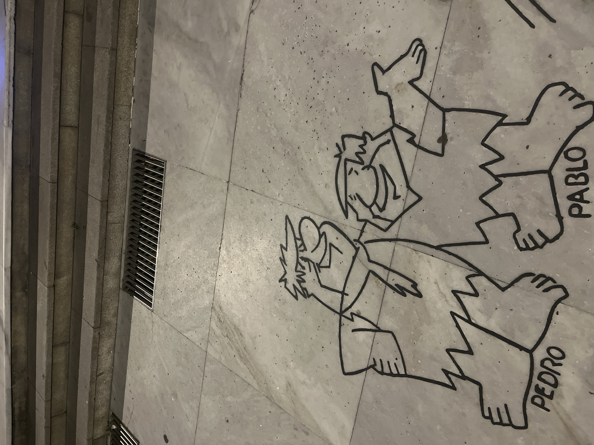 The image shows a street art depiction of characters from The Flintstones, specifically Pedro and Pablo, drawn in black on a concrete pavement. The characters are depicted in a simplified, outline style, with jagged edges on their clothing and feet. The word "PEDRO" is visible below the left character, and "PABLO" is visible below the right character. The pavement is segmented by grout lines, and in the background, there are stone steps leading upwards and a metal grate for drainage. This street art is located on Calle de la Marineda in A Coruña, Spain, near the Domus museum. The scene is outdoors, and the lighting suggests it is nighttime or a heavily shaded area.