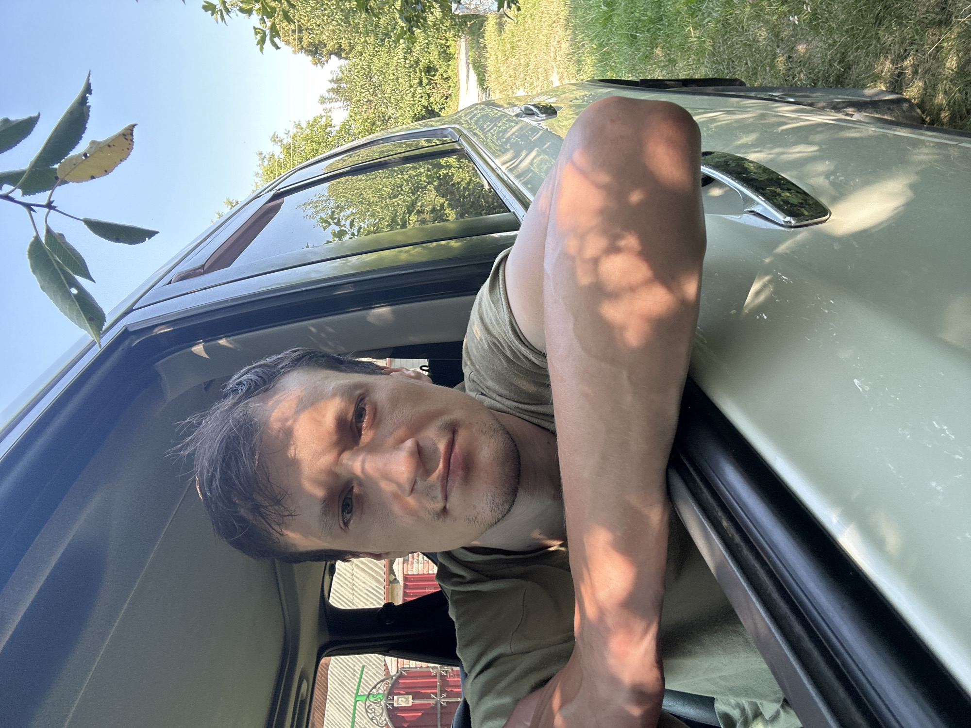 A man is leaning out of the open driver's side window of a car, his arms resting on the door frame. He is looking directly at the camera. The man has short, dark hair and is wearing a green t-shirt. Sunlight casts shadows of leaves and branches across his arms and face. In the background, trees and grass are visible, along with a fence and a building with a corrugated roof, consistent with the rural landscape of Shatava, Ukraine. The sky is clear and blue.