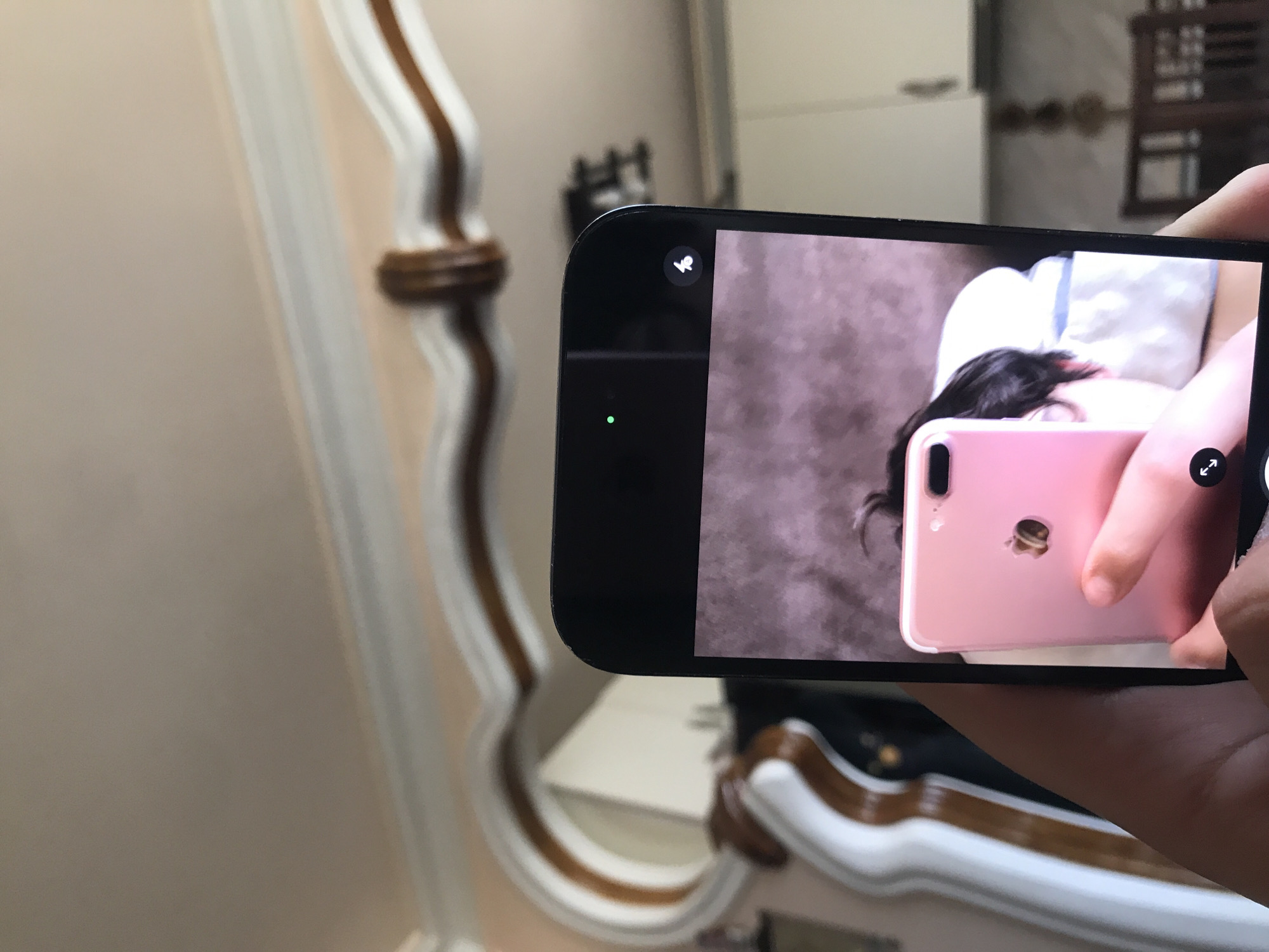 A person is holding a black smartphone to take a picture of a pink smartphone. The pink smartphone appears to be taking a selfie with a subject who has dark hair and is lying down. The background is blurry and shows a room with furniture and a mirror. The photo was likely taken in an indoor setting, and the location cannot be determined from the image.