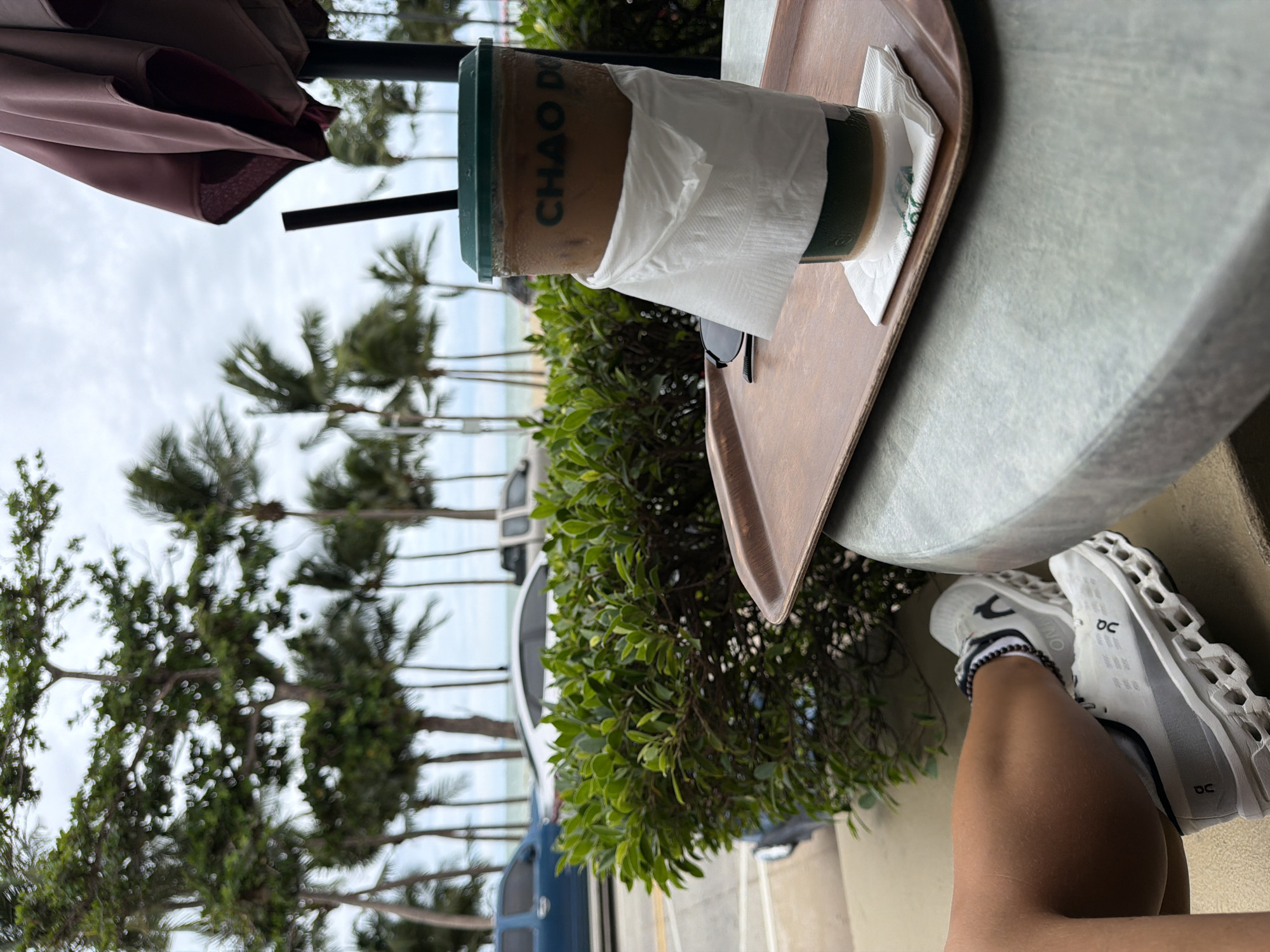 The scene captures an outdoor setting in Pattaya City, Thailand, with a focus on an individual's leisure experience by the coast. In the foreground, a person's lower leg and foot, clad in a white On Cloud athletic shoe and a black beaded anklet, are visible on the left. A light grey, textured table supports a brown wooden tray. On this tray, a clear plastic cup filled with iced coffee, wrapped in a white paper napkin, is positioned. The cup's green lid features a black straw, and the text "CHAO DO" is visible in green on its side. A pair of dark sunglasses also rests on the tray. To the right, a folded brown umbrella, typical of outdoor seating, is partially visible against an overcast sky.
The mid-ground features a vibrant green bush acting as a visual barrier. Beyond the bush, a paved road with vehicles, including a blue pickup truck, a white sedan, and a light-colored pickup truck, runs parallel to the coastline. In the background, a sandy beach transitions into the calm sea under a cloudy sky. The entire horizon is lined with numerous tall palm trees, characteristic of a tropical beachfront environment. The coffee brand and the coastal landscape strongly indicate a location such as Pattaya Beach Road or Jomtien Beach Road in Pattaya City.