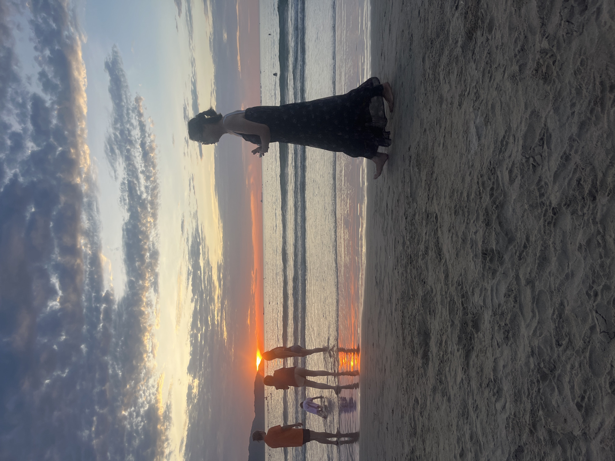 This image depicts a sunset scene on a beach in Đà Nẵng, Vietnam. The sun, a prominent orange-red orb, is visible just above the horizon line in the central left portion of the frame, casting intense light across the water and sky. Distant mountains or landmasses are silhouetted along the horizon.
The sky exhibits layered clouds: darker, denser formations occupy the upper frame, transitioning to lighter, more scattered clouds illuminated by the sunset's warm yellow and orange hues closer to the horizon. The ocean surface displays gentle waves breaking on the shore, with the shallow water reflecting the vibrant sunset colors. Several small, indistinct vessels are visible as dark silhouettes on the distant sea.
On the left side of the image, four individuals are positioned at the water's edge. One person, wearing an orange shirt and dark shorts, stands with their back mostly to the viewer. Another person, in a dark top and light shorts, faces the water, appearing to hold an object. A third individual, also in dark and light attire, stands further into the water. A child, dressed in light clothing, is on hands and knees in the shallow water, facing away from the viewer. Reflections of these individuals are visible on the wet sand.
On the right side of the image, a solo adult figure, appearing female, walks barefoot from right to left across the wet sand. This individual has dark hair tied back, wears glasses, and a long, dark, possibly patterned dress. They hold a small, light-colored object in their right hand. Their profile is presented, and they are silhouetted against the twilight sky.
The foreground consists of dark, wet sand with visible texture and subtle undulations. The overall lighting is characterized by the low, warm glow of the setting sun, creating strong backlighting and prominent reflections on the water.