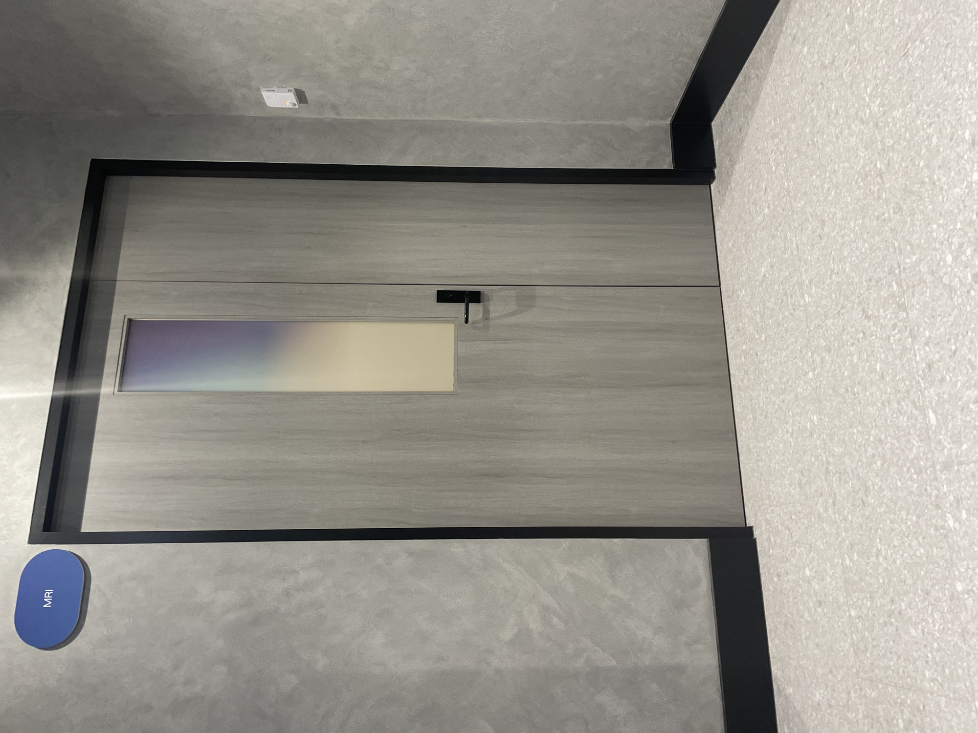 An oval, blue sign with the word "MRI" in white lettering is affixed to a grey, textured wall on the left side of the image. To the right of the sign is a closed, light grey wooden door with a black frame. The door has a rectangular frosted glass window that displays a gradient of iridescent colors. A black door handle is visible on the right side of the door. A small white electronic device is mounted on the grey wall to the right of the door. The floor is covered in light-colored terrazzo tiles. The scene appears to be indoors, possibly a hallway in a medical facility, with a contemporary and clean aesthetic. The location is not specified as Melbourne, Australia.