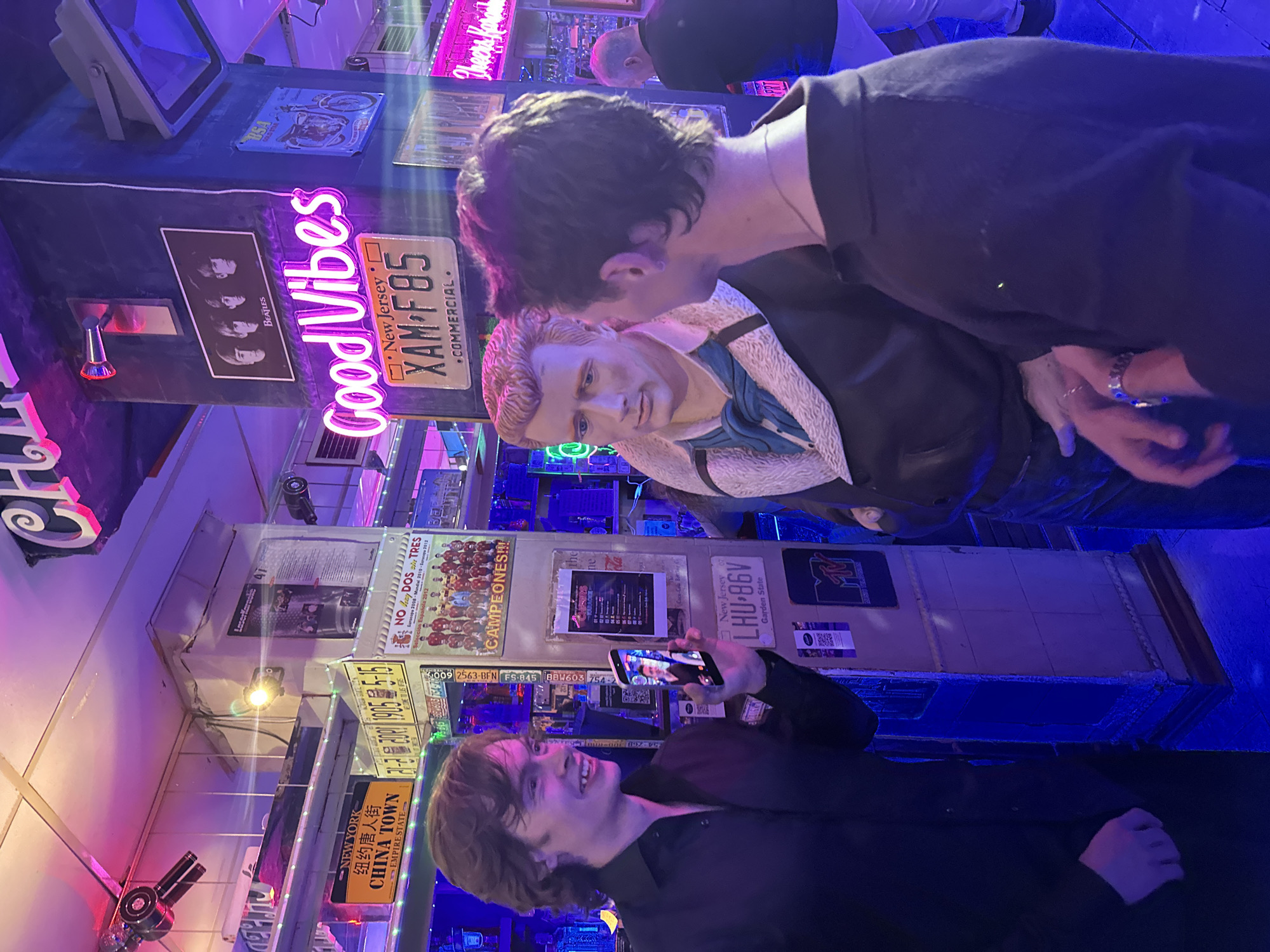 The image depicts the interior of a bar or entertainment venue. The walls are adorned with a collection of license plates, posters, and neon signs, including one that reads "Good Vibes" and a photo of The Beatles. A wax figure resembling Elvis Presley is a prominent feature. Two individuals are interacting with the display. One person, wearing a black jacket, is taking a photo with a smartphone. The other individual, also wearing dark clothing, appears to be interacting with the Elvis wax figure. The lighting is predominantly blue and purple, which contributes to the scene's ambiance. A license plate referencing "New York China Town" suggests a US theme. "Cheers Karaoke" is also visible in neon. The text "Laserkaraolic A" and "LaserKaraolic A" is partially visible. Based on the text "Cheers Karaoke" and "LaserKaraolic A", the image is likely taken in a Karaoke bar. This bar is located in Madrid, Spain.