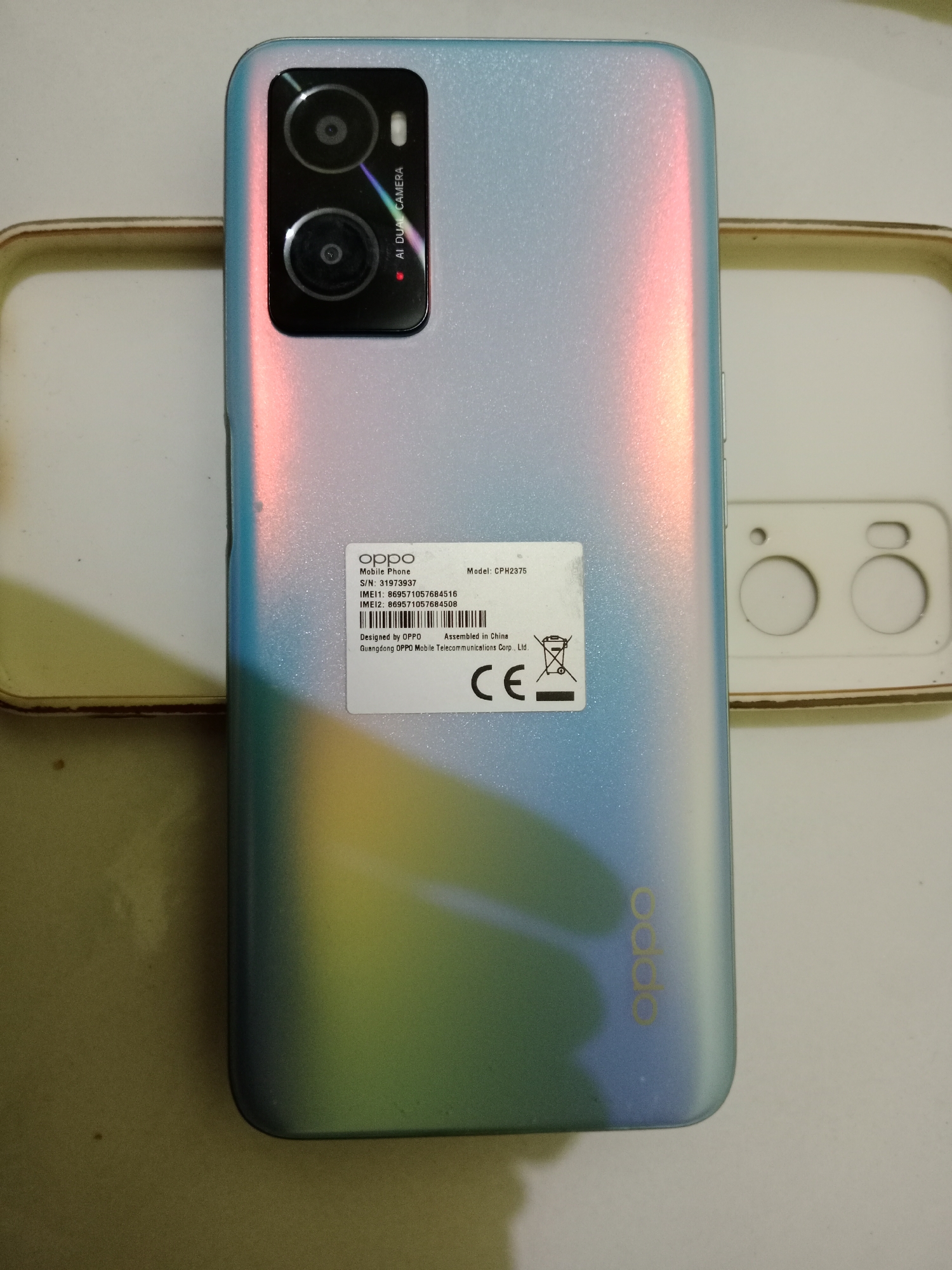A light blue and pink iridescent back of an Oppo smartphone is the primary subject, positioned diagonally on a white surface. The phone exhibits a dual-camera system on the top left, with text indicating "AI DUAL CAMERA." A white sticker on the phone's back contains manufacturing details, including "OPPO Mobile Phone," "S/N: 31973937," "IMEI1: 869571057684516," "IMEI2: 869571057684508," "Model: CPH2375," "Designed by OPPO," "Assembled in China," and "Guangdong OPPO Mobile Telecommunications Corp., Ltd." A recycling symbol and "CE" mark are also present. The Oppo logo is visible in gold lettering on the lower right side of the phone. To the left of the phone, a light brown phone case is partially visible. To the right, a white plastic frame, possibly for packaging or display, with two circular cutouts for camera lenses, is also present. The lighting creates a gradient of colors on the phone's surface, from pink on the left to blue and green on the right. The image does not contain any identifiable elements indicating its location in West Jakarta, Indonesia.