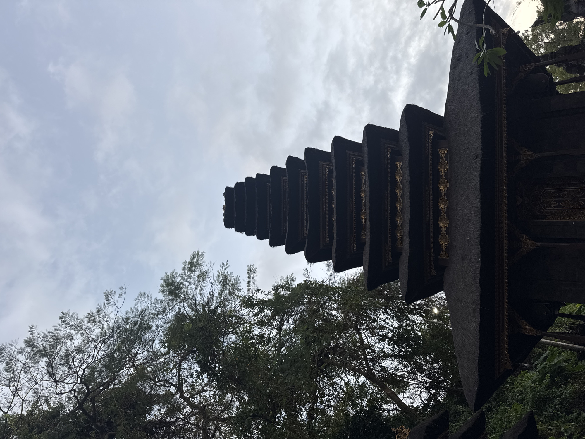 An upward-angled, low-angle full shot captures a multi-tiered, dark pagoda-style temple structure with thatched roofing and ornate gold trim. The temple is set against a cloudy, grey sky, with lush green trees and foliage visible to the left and right of the structure. The scene is likely Dawan, Indonesia, given the architectural style and vegetation, possibly representing a Hindu temple. The lighting is dim and diffused, suggesting an overcast day or early morning/late evening. No people are present in the frame.