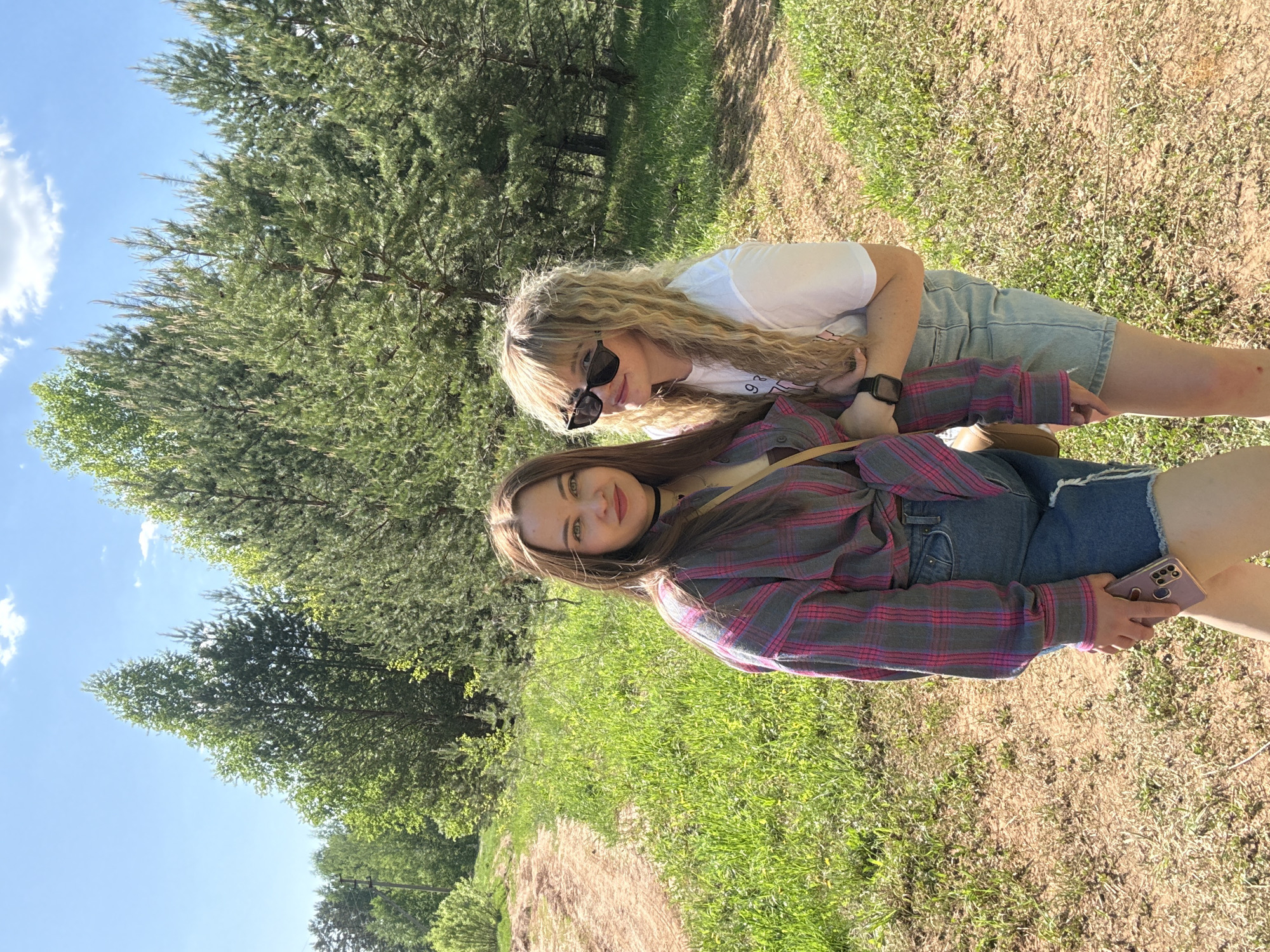 Two women are in an outdoor setting. The foreground is characterized by grassy and sandy ground, suggesting a field or trail. The woman in the foreground is wearing a pink and gray plaid shirt and denim shorts, and holding a cellphone. The other woman is wearing a white shirt and denim shorts, and sunglasses. Both women are close together, likely posing for the photo. The background is dominated by tall green trees under a blue sky. The image appears to be taken in a natural environment, likely in the city of Марковский, Russia.