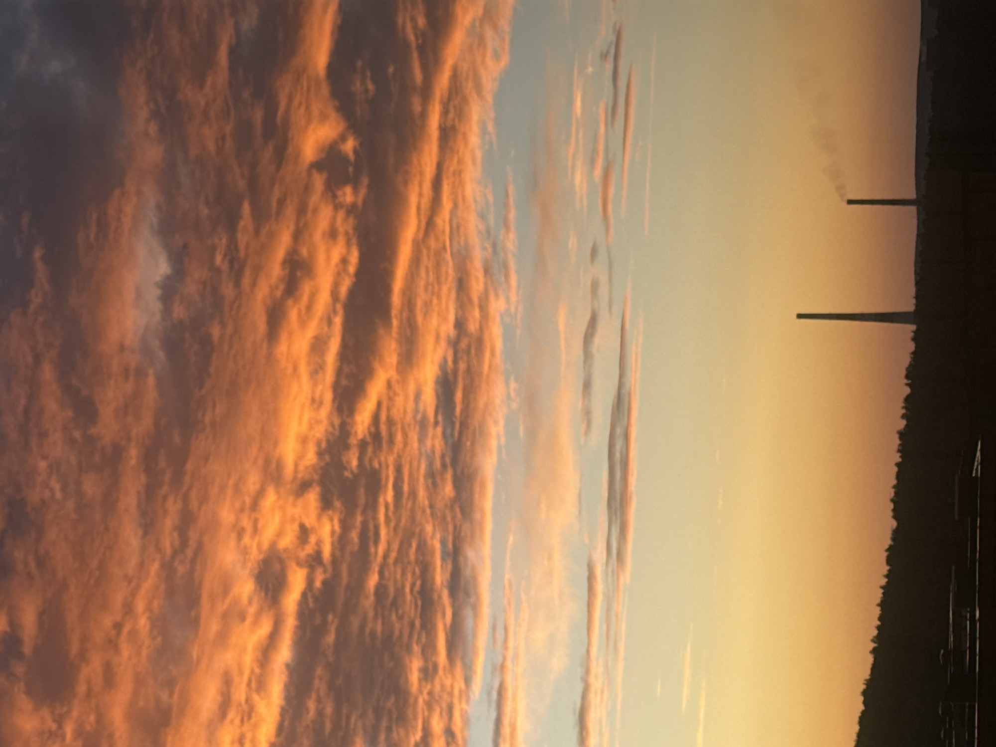 The image captures a sunset over the city of Markova, Russia. The sky is dominated by vibrant orange and pink clouds, with patches of lighter blue and yellow. In the foreground, a dark silhouette of a tree-covered hill runs across the bottom of the frame. Two industrial smokestacks are visible against the horizon; one is taller and emitting a plume of smoke, while the other is shorter and also shows faint smoke. The landscape below the hill appears to be industrial buildings, silhouetted against the warm sky.
