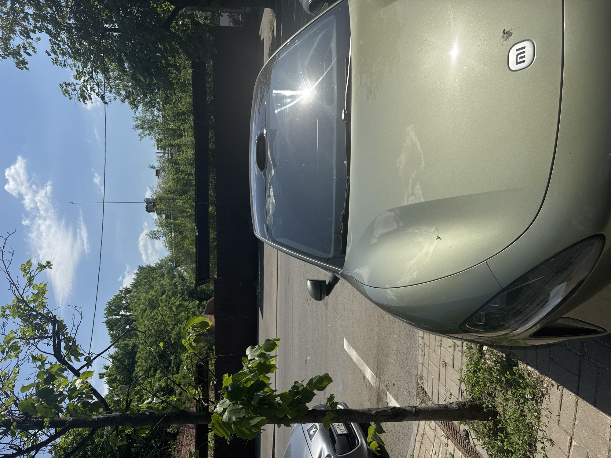 The image depicts a light green electric car parked on a paved street next to a tree in Kazan, Russia. The car has an "E" logo on the right rear panel. Sunlight glares off the windshield. A dark brown wooden fence runs along the left side of the frame, behind which are buildings, trees, and a blue sky with clouds. A telephone pole is visible in the background. Another silver car is partially visible under the tree. The ground is paved.