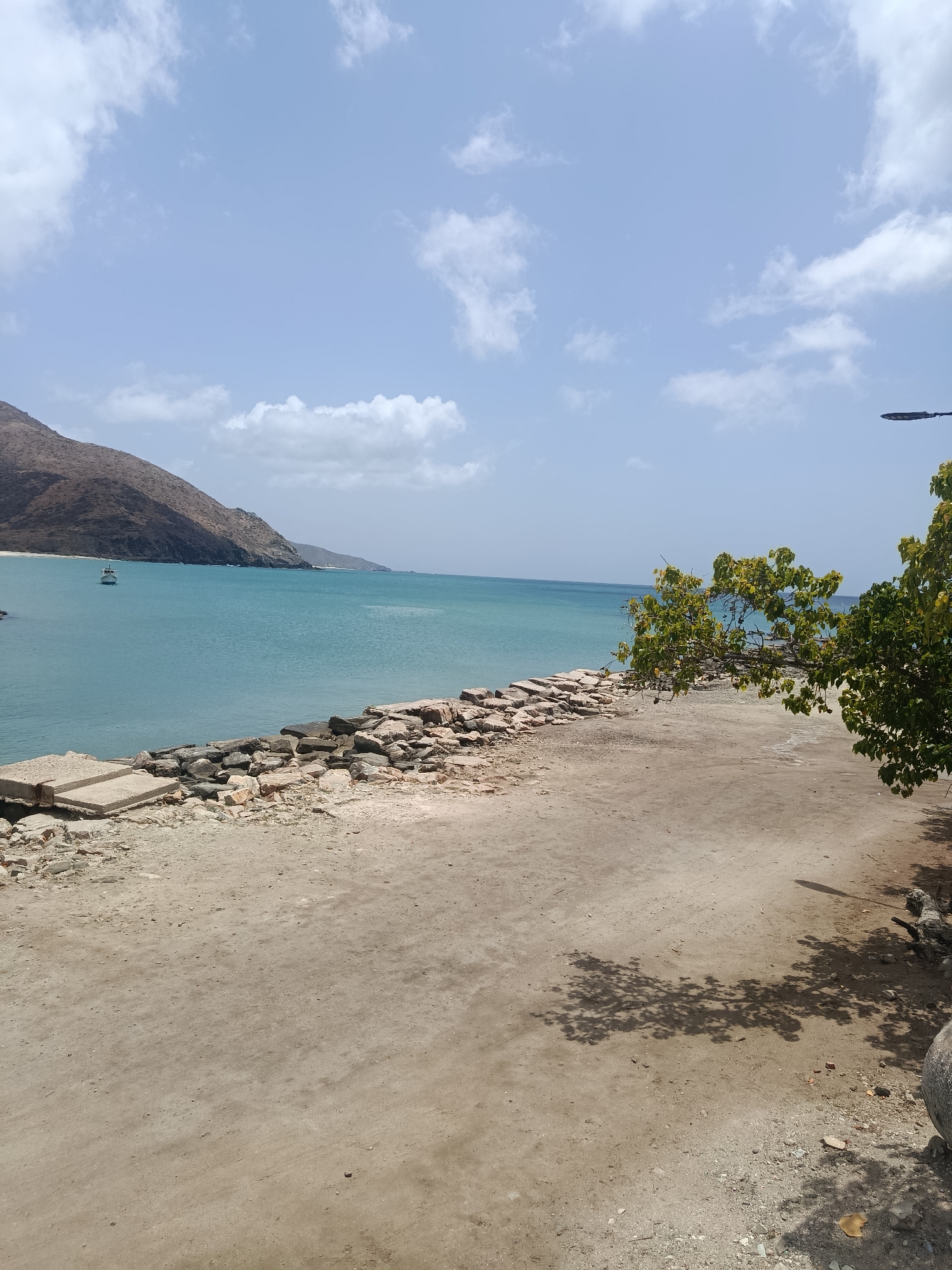 The scene depicts a coastal area in Pedro González, Venezuela. The foreground is a sandy, dirt-covered area with some exposed rocks and debris. A low stone wall, constructed of concrete and loose rocks, separates this area from the turquoise sea. In the sea, a small boat is visible in the distance. A mountainous landmass is visible on the left side of the frame. A tree with green foliage is located on the right side, casting shadows on the ground. The sky is blue with scattered white clouds. A street light pole is visible on the right side.