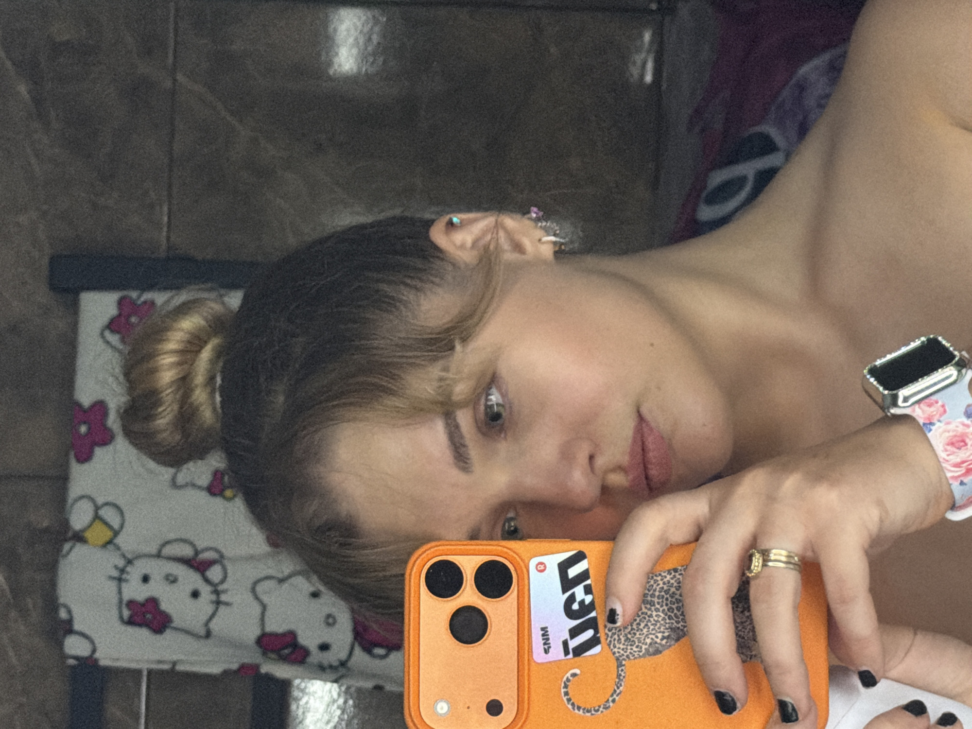 A close-up, rotated selfie captures a young woman's reflection in what appears to be a mirror or glass surface, taken indoors in Большая Шильна, Russia. The woman, with light brown hair styled in a bun and bangs, looks directly towards the camera. Her right ear is visible with multiple piercings adorned with small, colorful studs.
She is wearing a sleeveless top or bare-shouldered attire, revealing her collarbone and shoulder. On her right wrist, she wears a smartwatch with a decorative white band featuring pink and blue floral patterns. A gold ring with a central stone or design is visible on her right ring finger. Her fingernails are painted with dark polish.
The woman holds an orange smartphone with a triple camera lens array in her right hand. The phone case features a sticker with a leopard print design and partially visible reversed text: "NGJu" and "MNS", along with a red "R" logo. A white blanket with Hello Kitty characters and pink flowers is visible beneath her head, suggesting a casual, home environment. The background consists of a dark, mottled, possibly tiled wall. The overall scene depicts a casual selfie being taken.