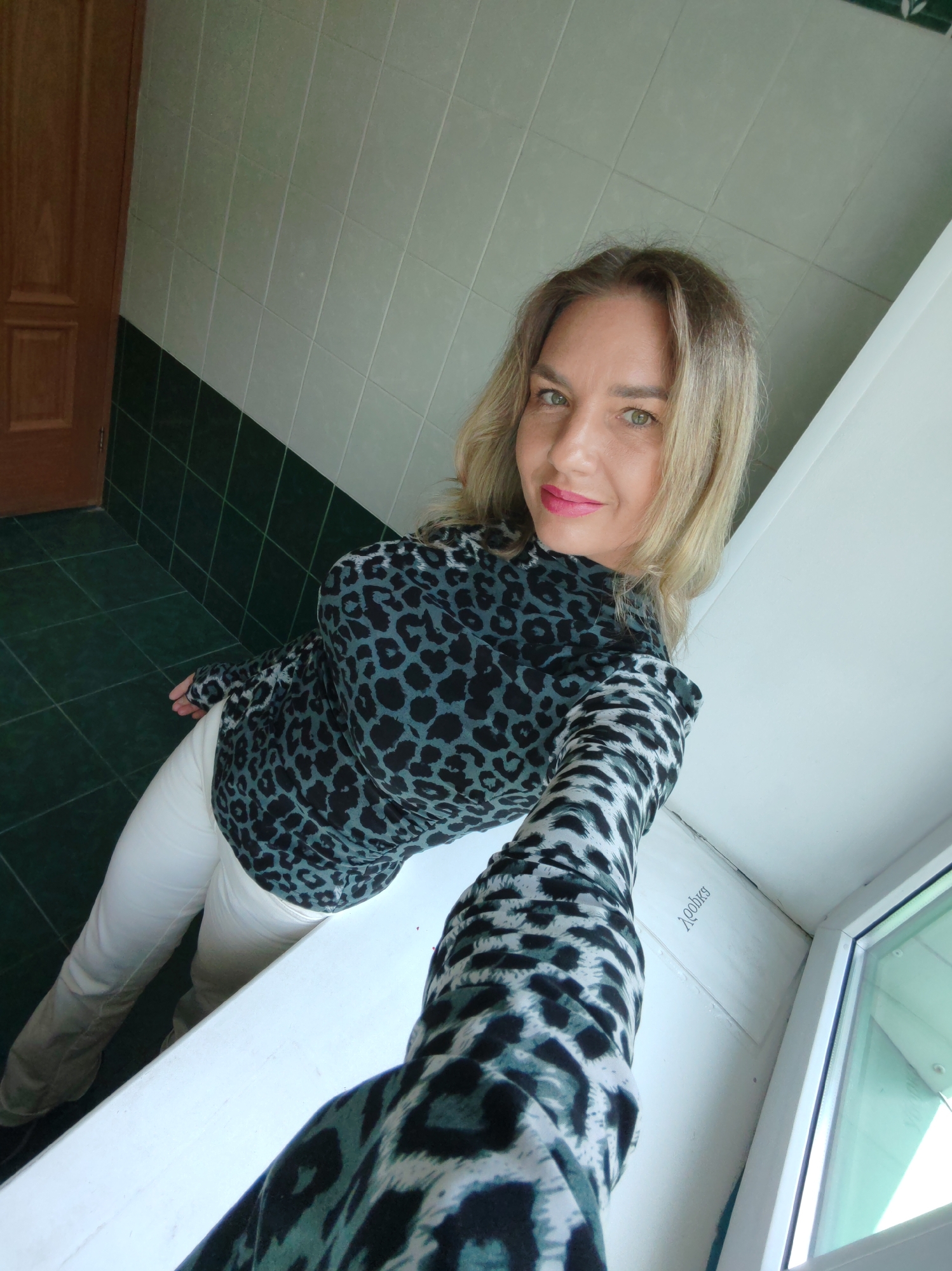 A woman with blonde hair and green eyes is captured in a selfie-style photograph. She is looking directly at the camera with a subtle smile and bright pink lipstick. She wears a long-sleeved top featuring a black and teal leopard print pattern, paired with light-colored (possibly white or off-white) trousers. Her arm, also clad in the leopard print sleeve, extends towards the right of the frame, indicating she is holding the camera.
The setting appears to be an indoor space with tiled walls and floor. The lower section of the wall is covered in dark green square tiles, while the upper section has lighter, possibly greyish-green, rectangular tiles. The floor is also tiled with dark green squares. A dark brown wooden door with a panel design is visible on the left side of the image. To the right, a white window sill or ledge runs across the frame, leading to a window with a white frame and a slight green tint to the glass.
The photograph is taken from a slightly elevated, downward-angled perspective, characteristic of a handheld selfie. The lighting is bright and even, without harsh shadows. On the white surface near the window, faint text is visible, which appears to read "joshea" or "ioshka". The overall mood conveyed by the woman's expression is pleasant and calm.