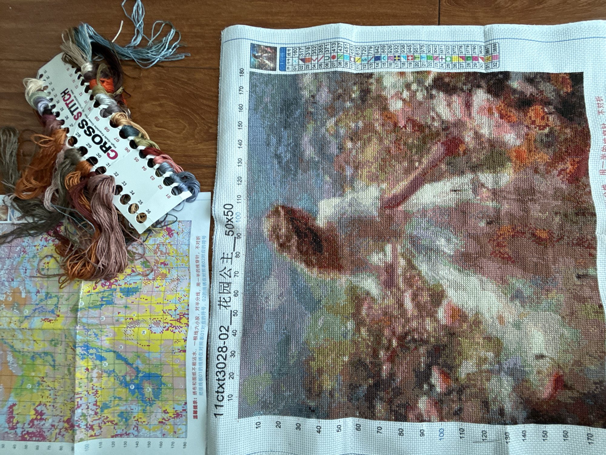 The image displays a close-up view of a cross-stitch embroidery kit. In the foreground, a partially completed cross-stitch canvas shows an image of a woman in a white dress with a floral background. The canvas has a grid pattern with numerical markings along the sides indicating stitch counts. To the right of the canvas is a color guide with numbered threads, each representing a specific color used in the embroidery. A tangled collection of embroidery threads is spread across the top and right side of the image, predominantly in shades of brown, pink, and blue. Some of the threads are attached to a white card labeled "CROSS STITCH" with corresponding numbers. The background consists of a wooden surface. The image is likely taken in a home or crafting environment. There is no indication of the city of Khan Mean Chey, Cambodia, in the image.