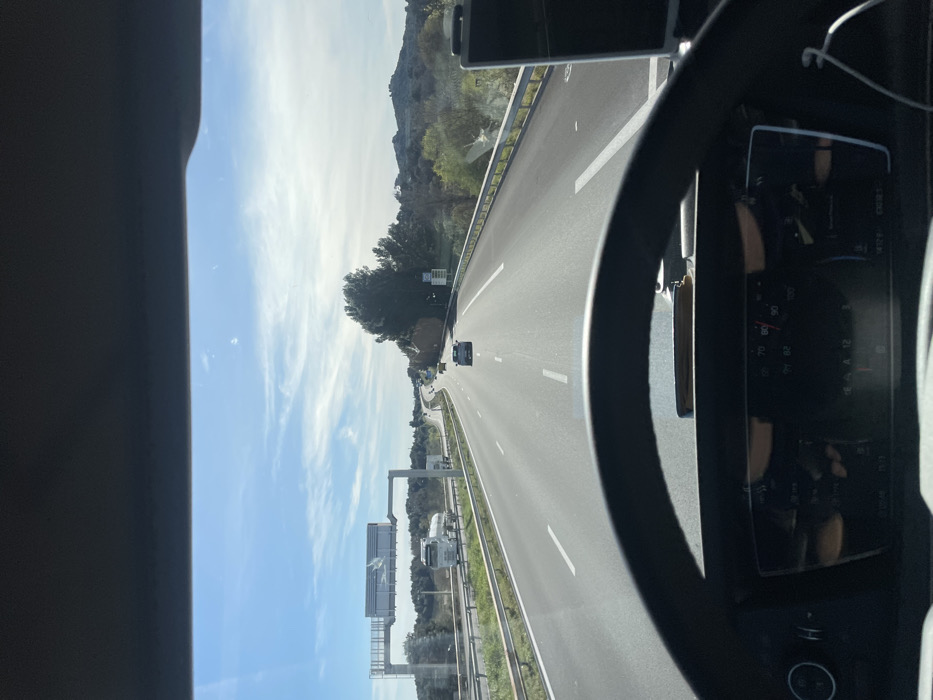 The image depicts a forward-facing view from inside a vehicle traveling on a multi-lane highway in Tavel, France, under a clear blue sky with scattered white clouds. The lower third of the frame is occupied by a dark-colored steering wheel and a portion of the vehicle's digital instrument cluster, which displays a speed of approximately 70-80 km/h. A dark object, possibly a wallet, rests on the dashboard in the center. To the right of the steering wheel, a dark electronic device, likely a smartphone, is mounted, with a white charging cable extending downward.
The highway is an asphalt road with white dashed lane markings, showing at least two lanes of travel in the depicted direction. Ahead in the right lane, a light-colored car is visible, followed by a darker vehicle further in the distance. In the left lane, a white tanker truck is also traveling forward. An overhead gantry structure spans the highway on the left, supporting several unreadable rectangular signs. Metal guardrails line both sides of the road. The environment surrounding the highway features green trees and bushes, with low hills visible on the horizon to the right. The scene is illuminated by bright daylight.