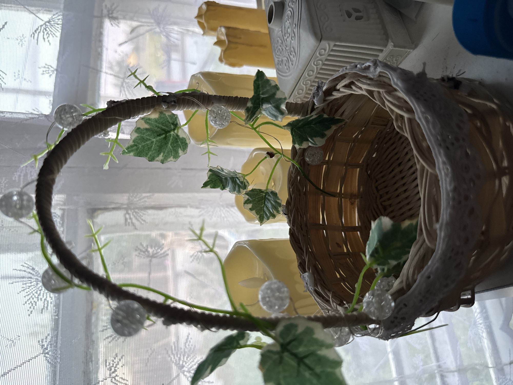A close-up, eye-level, medium shot captures a decorated wicker basket in the foreground, positioned on a surface, likely a shelf or windowsill, against a backdrop of sheer white curtains. The basket is woven from light-colored material and features a dark brown handle, also woven, that arches over the opening. Green artificial ivy with variegated leaves is intertwined with the handle, along with small, clear, faceted ornaments that resemble beads or small spheres. A delicate white lace trim edges the top of the basket.
Behind the basket, the sheer curtains have a subtle, almost snowflake-like pattern, suggesting a window. Beyond the curtains, the scene is blurred, but the shapes of at least three tall, cylindrical, yellow objects, possibly LED candles, are visible. To the right of the basket and candles, a rectangular, white decorative box with intricate embossed patterns sits on the same surface. A small, dark object, possibly a lid or a cap, rests on top of this box.
The overall impression is one of a decorative still life, possibly arranged for a festive occasion or as a display. The lighting appears soft, originating from the window, and the focus is sharpest on the basket and its embellishments. There are no people or animals present in the scene, and no identifiable actions are occurring. The environment suggests an indoor setting, possibly a home or a shop.
Regarding the location in Nyurba, Russia, there is no visual information in the image that allows for this specific geographical identification. The scene is generic and could be found in many locations.