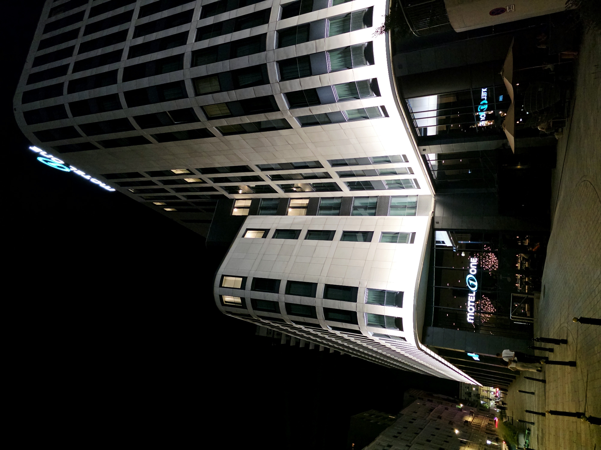 The image depicts the Motel One hotel in Warsaw, Poland, at night. The modern architecture of the hotel features a white facade with many windows, some illuminated from within. A neon sign spelling "Motel One" is visible on the building. The street in front of the hotel is paved with cobblestones and lined with bollards. Several people are walking on the sidewalk and seated at outdoor tables with umbrellas. The scene conveys a lively urban atmosphere at night.
