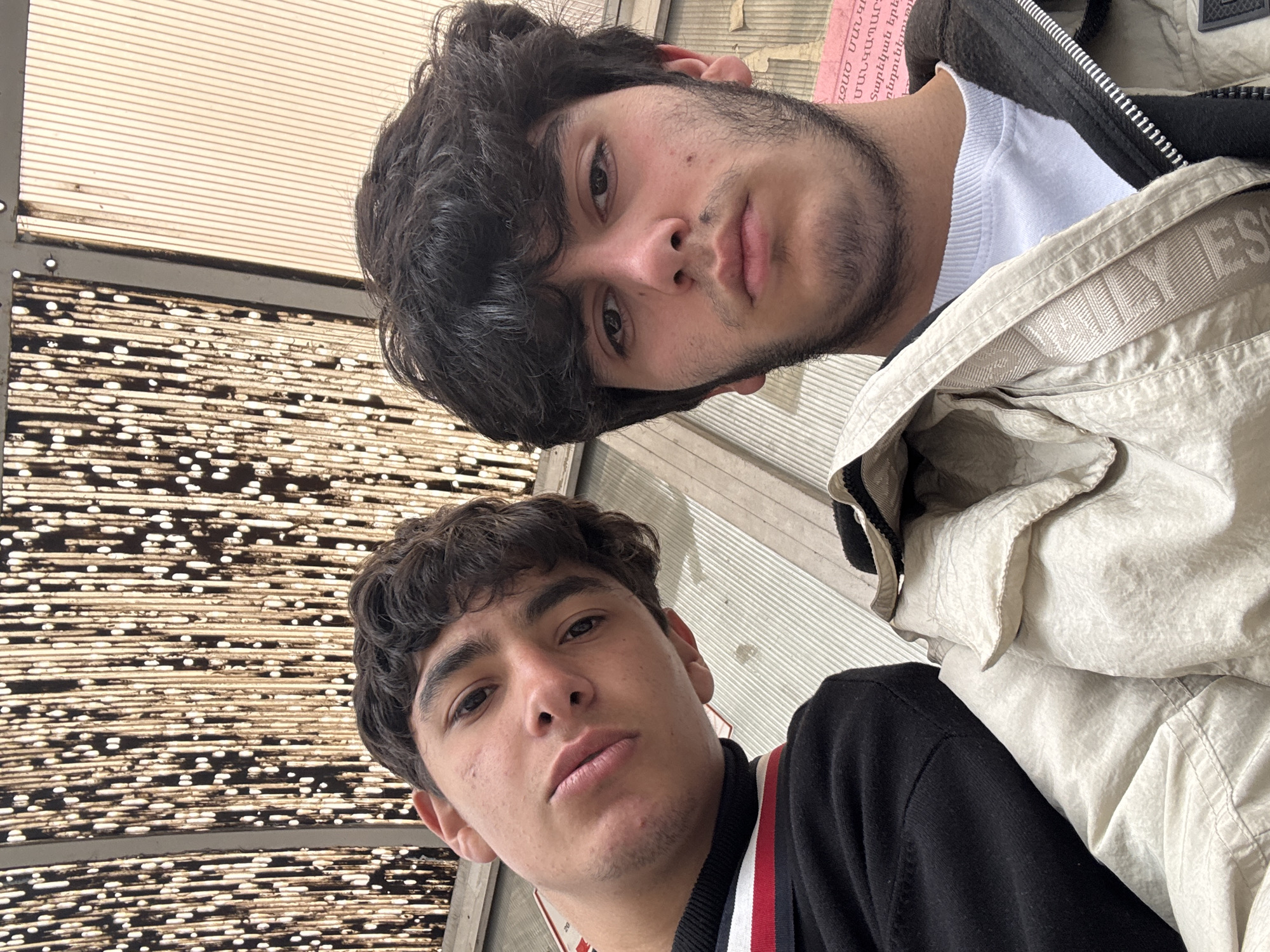 Two young men are posing for a selfie. The top man is wearing a white t-shirt under a black jacket, with a beige jacket partly covering him. The bottom man is wearing a black jacket with red, white, and blue stripes on the shoulder. The background features a ceiling or roof structure of horizontal metal slats, some with dark spots or holes. The image may have been taken in Yerevan, Armenia, given the presence of what appears to be Armenian text in the background on a pink surface partially visible between the two subjects.