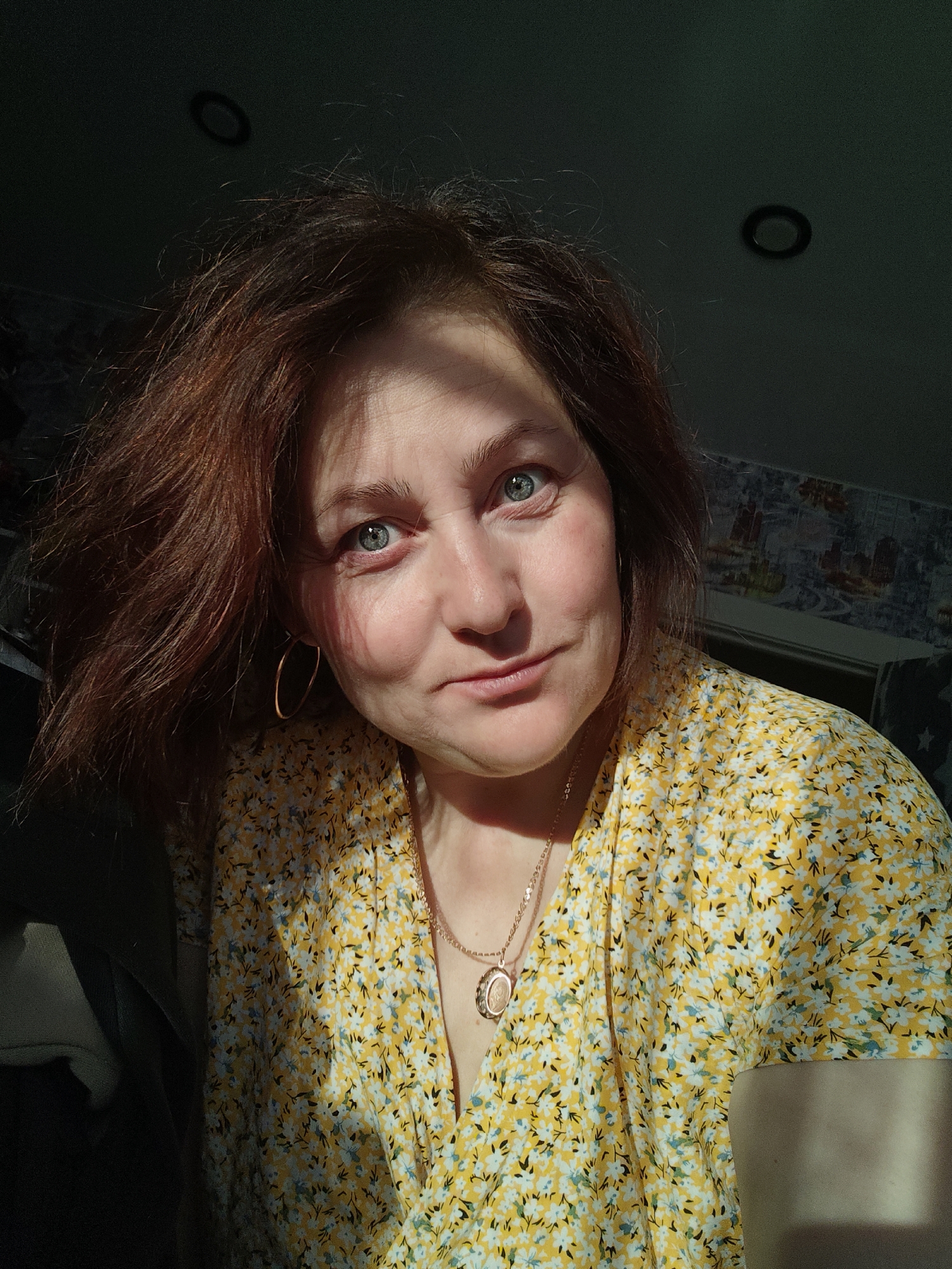 A woman with red hair and blue eyes is looking at the camera with a slight smile. She is wearing a yellow floral dress and a gold necklace with a pendant. She also has gold hoop earrings. The lighting is partially in shadow, highlighting one side of her face. Two ceiling lights are visible in the background. Based on details that cannot be extracted from the image, it is impossible to ascertain any specific details about the location of the image.