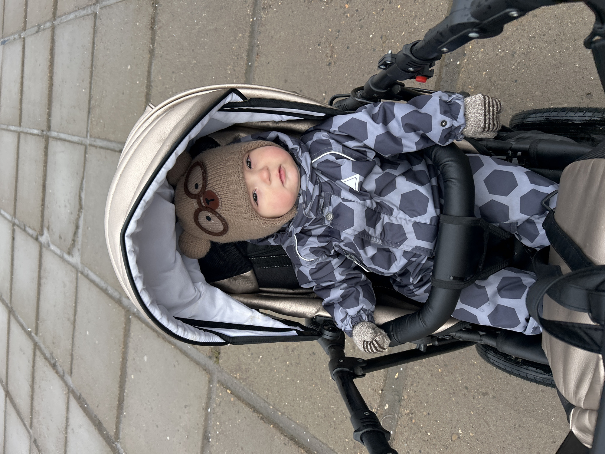 This media file captures a baby dressed in warm winter attire, seated in a stroller outdoors. The baby, an infant or young toddler, is wearing a light brown knitted hat adorned with features resembling a bear's ears and glasses. They are also clad in a gray and dark gray snowsuit with a distinct hexagonal or honeycomb pattern, along with light-colored, striped mittens. The baby looks directly at the camera with a calm, neutral expression.
The baby is positioned in a modern stroller or pram, characterized by cream or champagne-colored padded fabric and sturdy black frame elements. The background consists of a paved surface made of light gray concrete slabs, which appear somewhat damp or speckled, suggesting cold or wet conditions. The diffused lighting indicates an overcast day. The overall scene suggests an outdoor excursion, likely a walk, taking place in cold weather, consistent with the given location context of Chaikovsky, Russia. There is no visible text in the image.