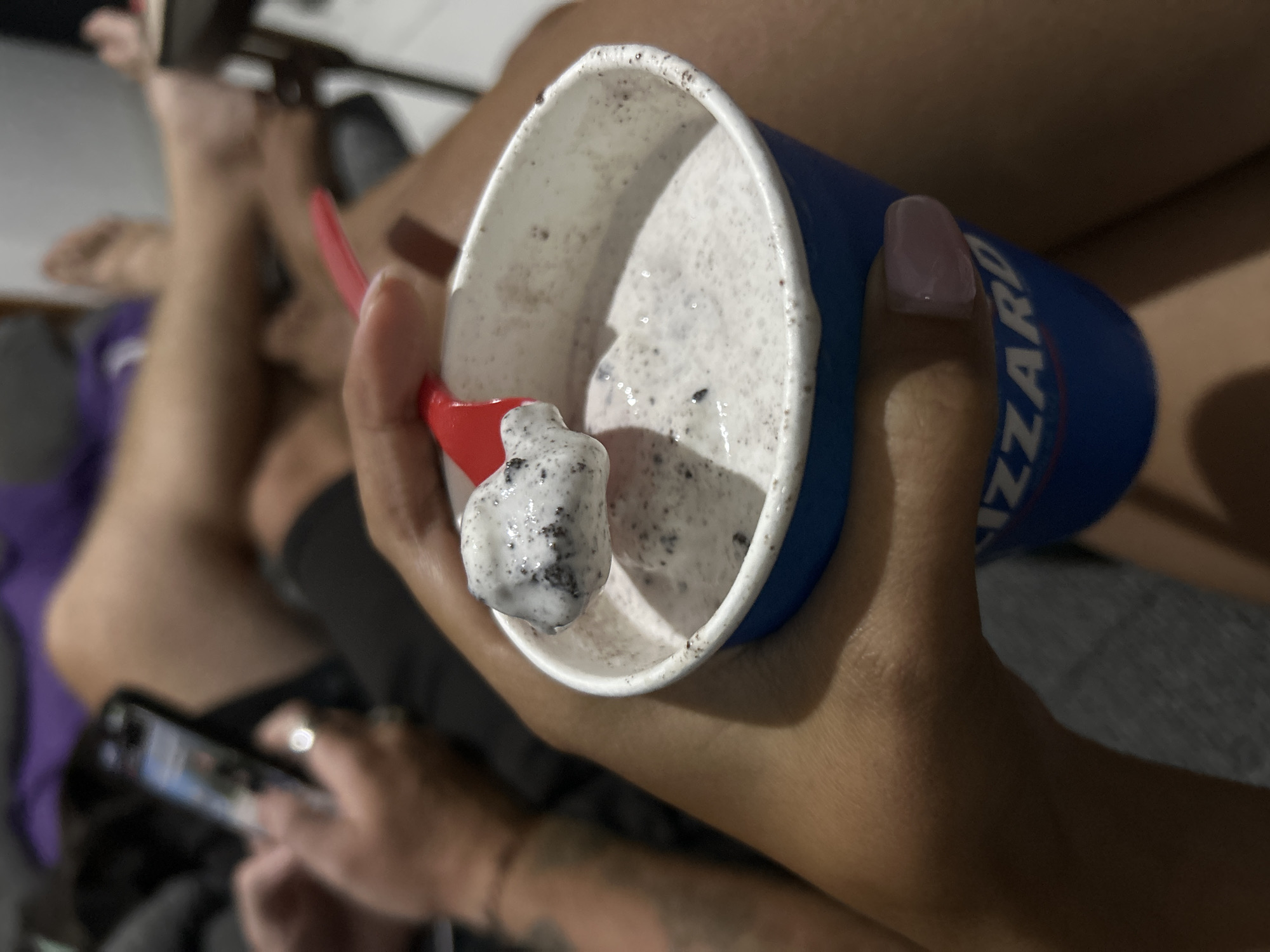 The image shows a close-up of a person holding a cup of cookies and cream ice cream with a red spoon. The ice cream is speckled with dark cookie pieces. The person's hand, with light pink nail polish, is holding the cup, and their thumb is visible resting on the blue outer layer of the cup. Partially visible in the background are other people's legs and feet, suggesting a casual gathering or outing. One person in the background appears to be looking at a smartphone. The overall scene appears to be indoors or in a shaded outdoor area, given the lighting. The text "BLIZZARD" is partially visible on the blue cup, indicating the brand of ice cream. The image evokes a sense of enjoying a sweet treat in a relaxed setting.